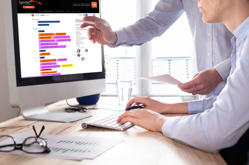 Two people work together at a desk, reviewing highlighted data on a computer screen and paper documents.
