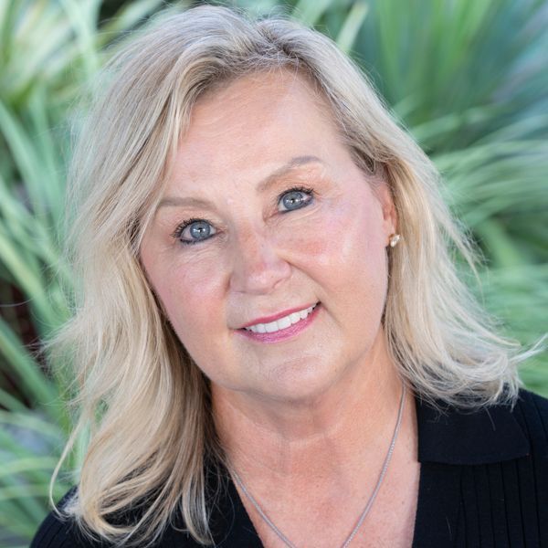 Blonde woman with blue eyes smiles, wearing black shirt, outdoors with blurred greenery.