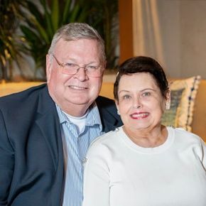 Man and woman smiling, indoors. Man in blue blazer, glasses. Woman in white sweater.