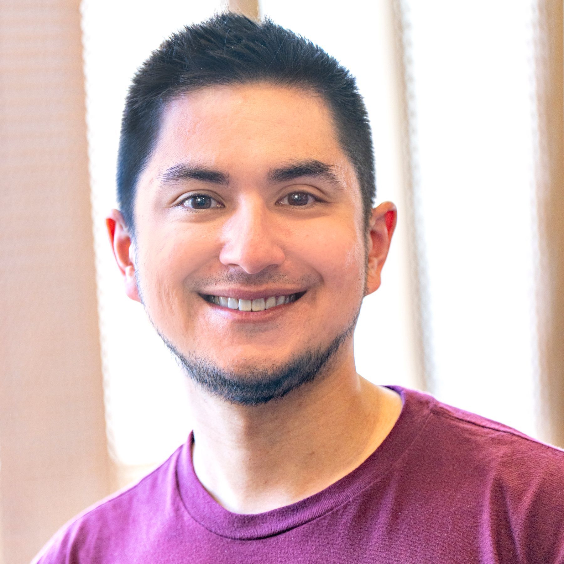 A young man (Jeremy Ott) with dark hair and a goatee wears a purple t-shirt and smiles at the camera.