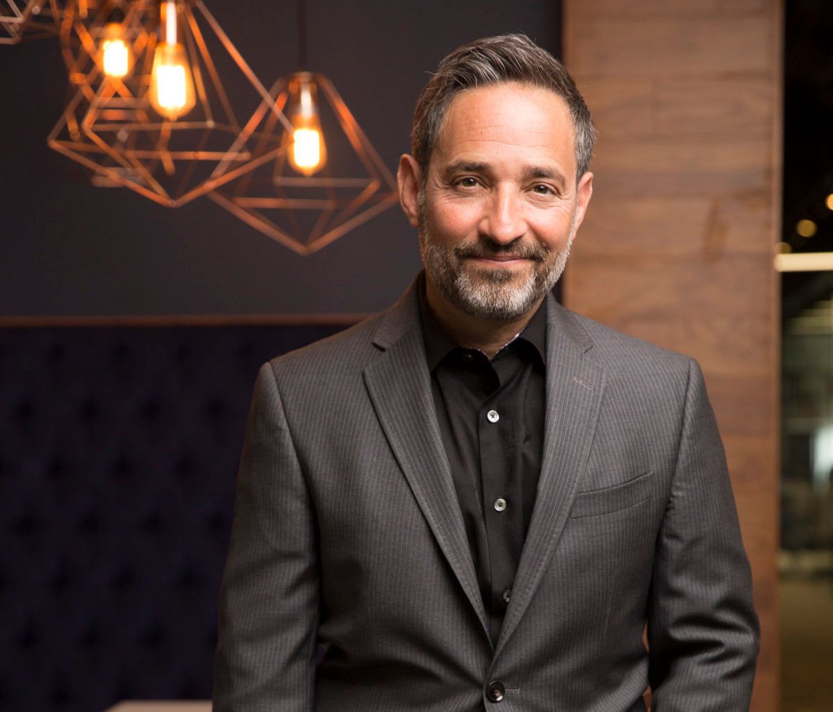 Man in blue sweater and pants smiles, leans against a window frame; city buildings visible outside.