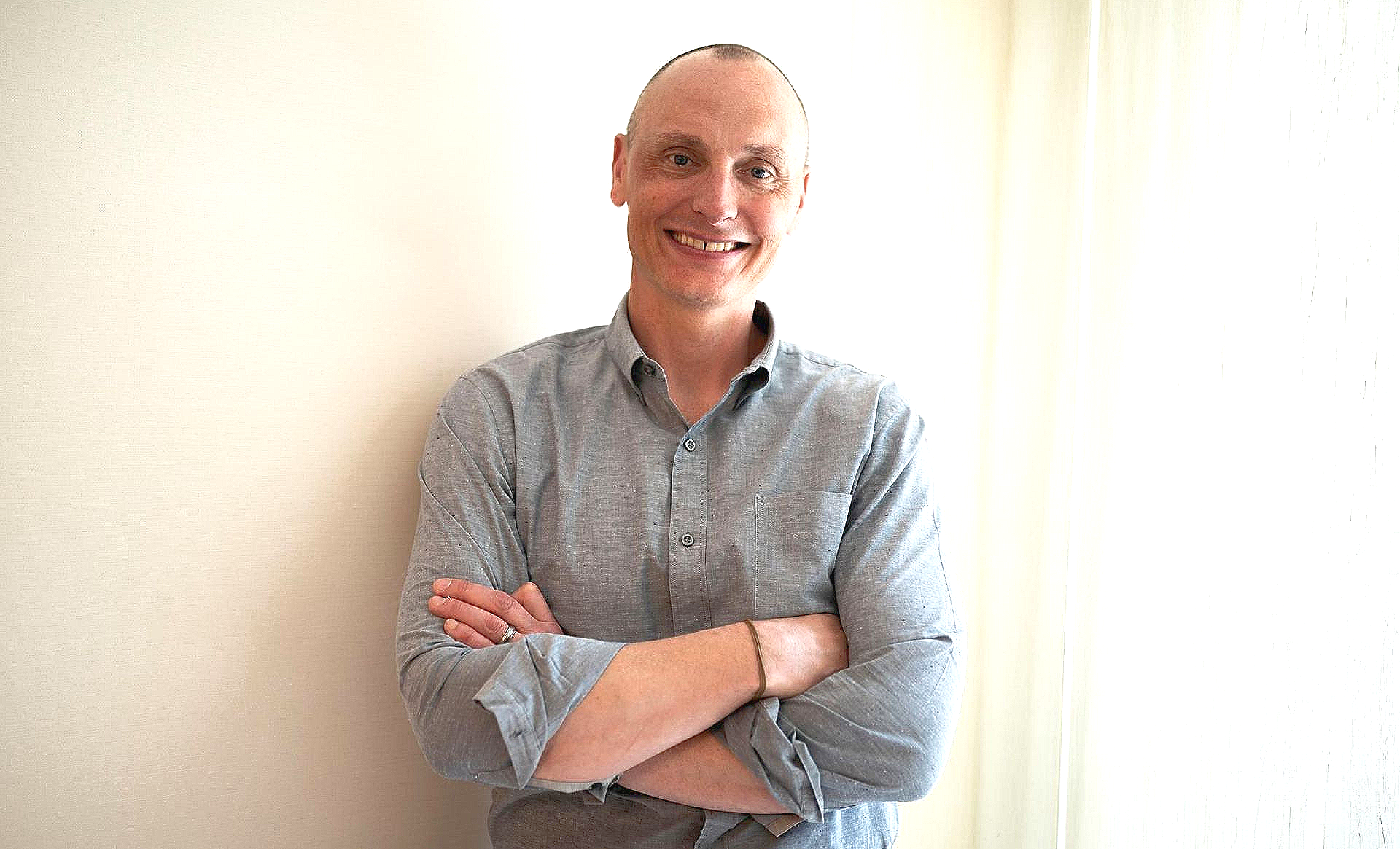 Man with crossed arms smiling, wearing a gray button-down shirt, leaning against a white wall.