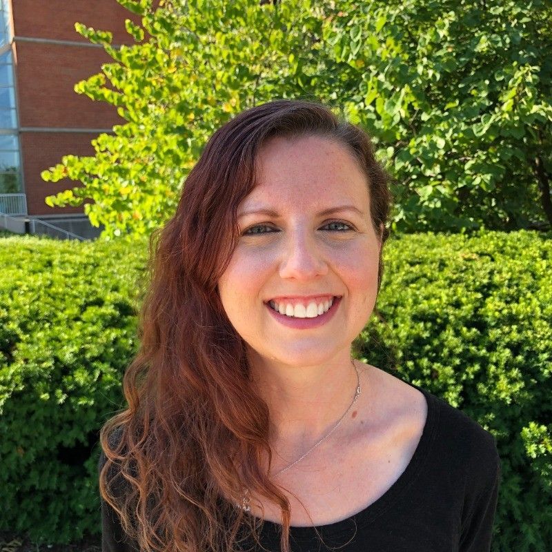 Woman with long reddish-brown hair smiles outdoors in front of green shrubs, wearing a black top.