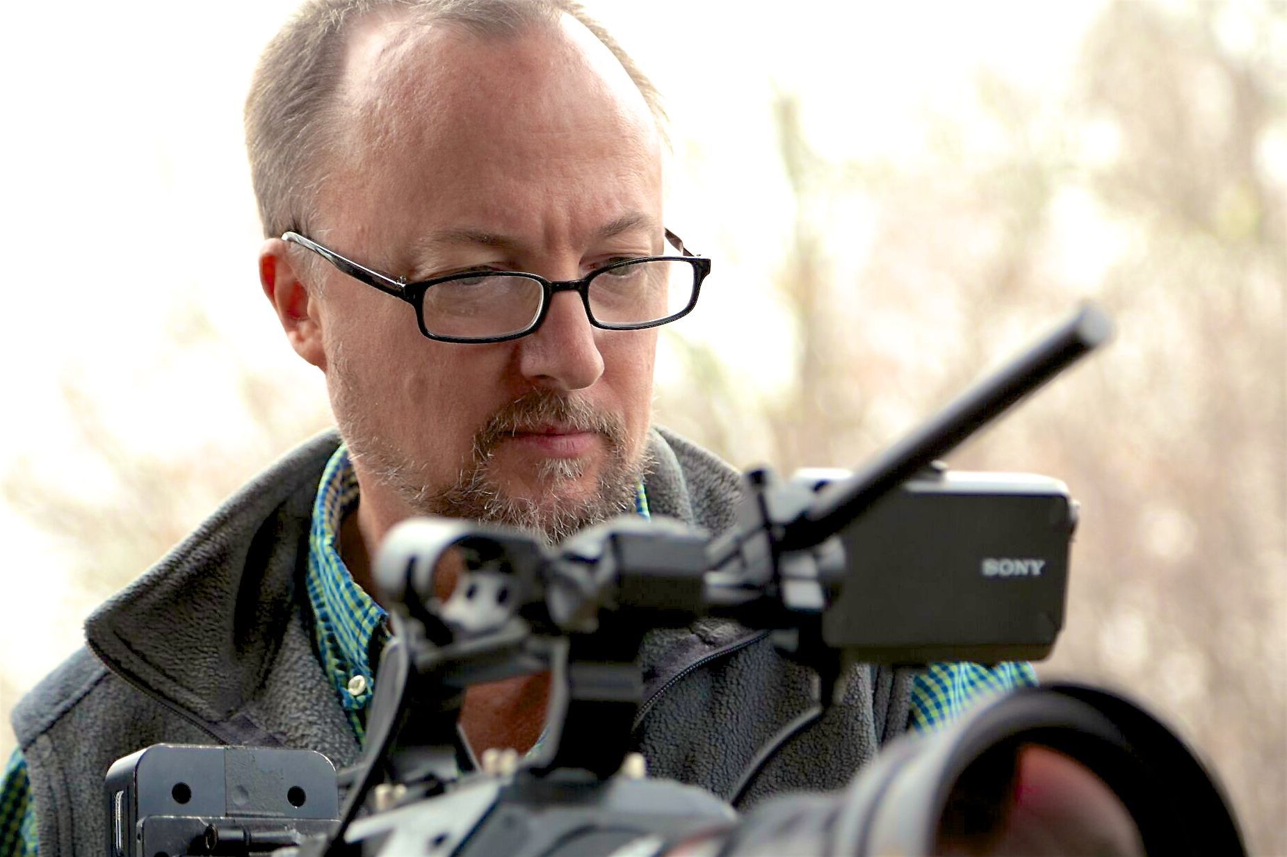 Man wearing glasses looking through a Sony video camera, outdoor setting.
