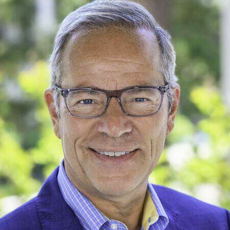 A graying man wearing purple glasses, and a purple suit, smiles at the camera.