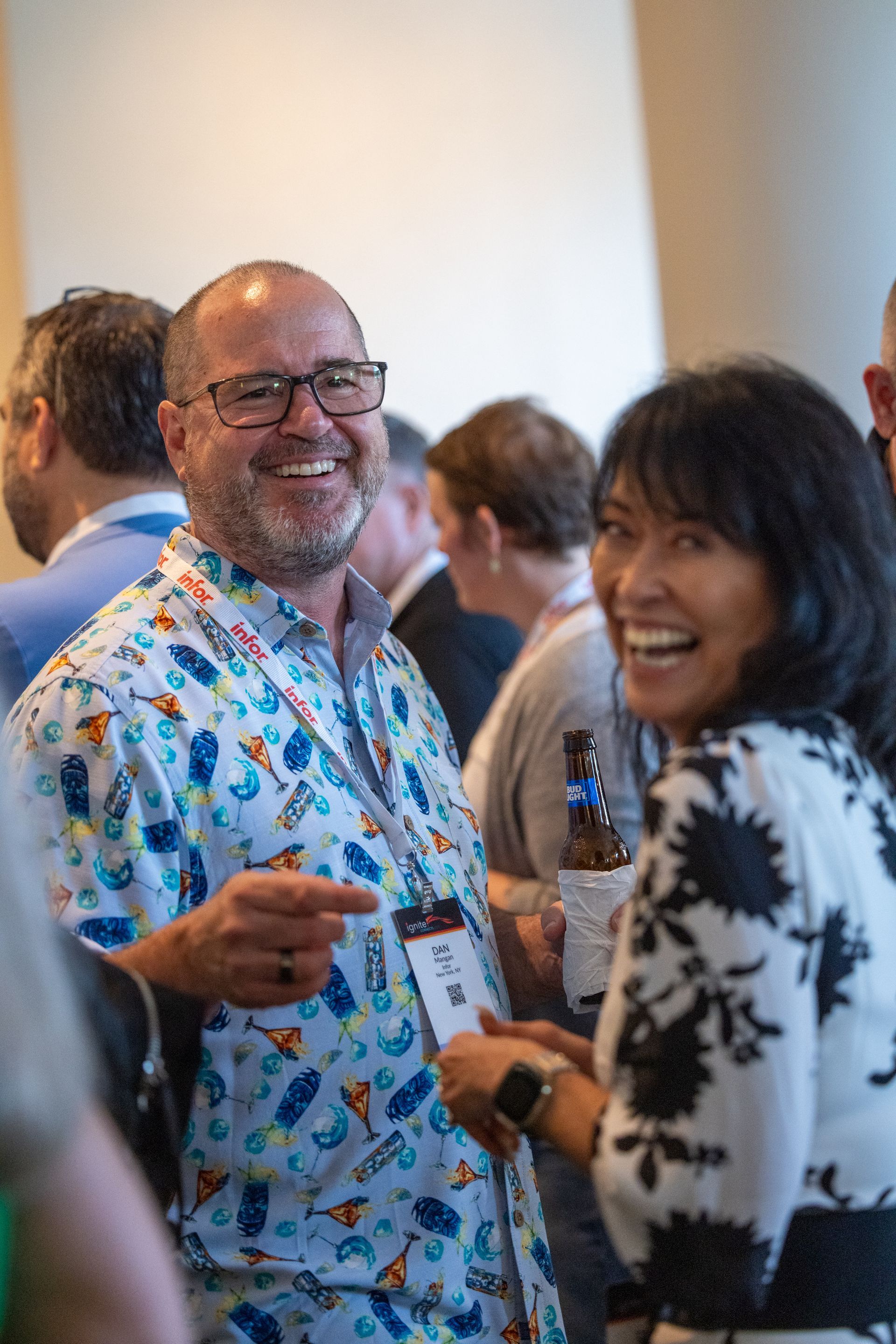 A man wearing a blue shirt with print of cocktail glasses and a woman wearing a white dress with a black floral print smile at the camera