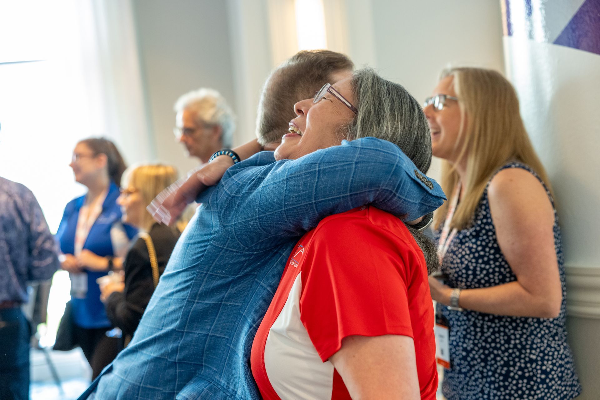 A person wearing a blue shirt hugs a woman in a white and red shirt.