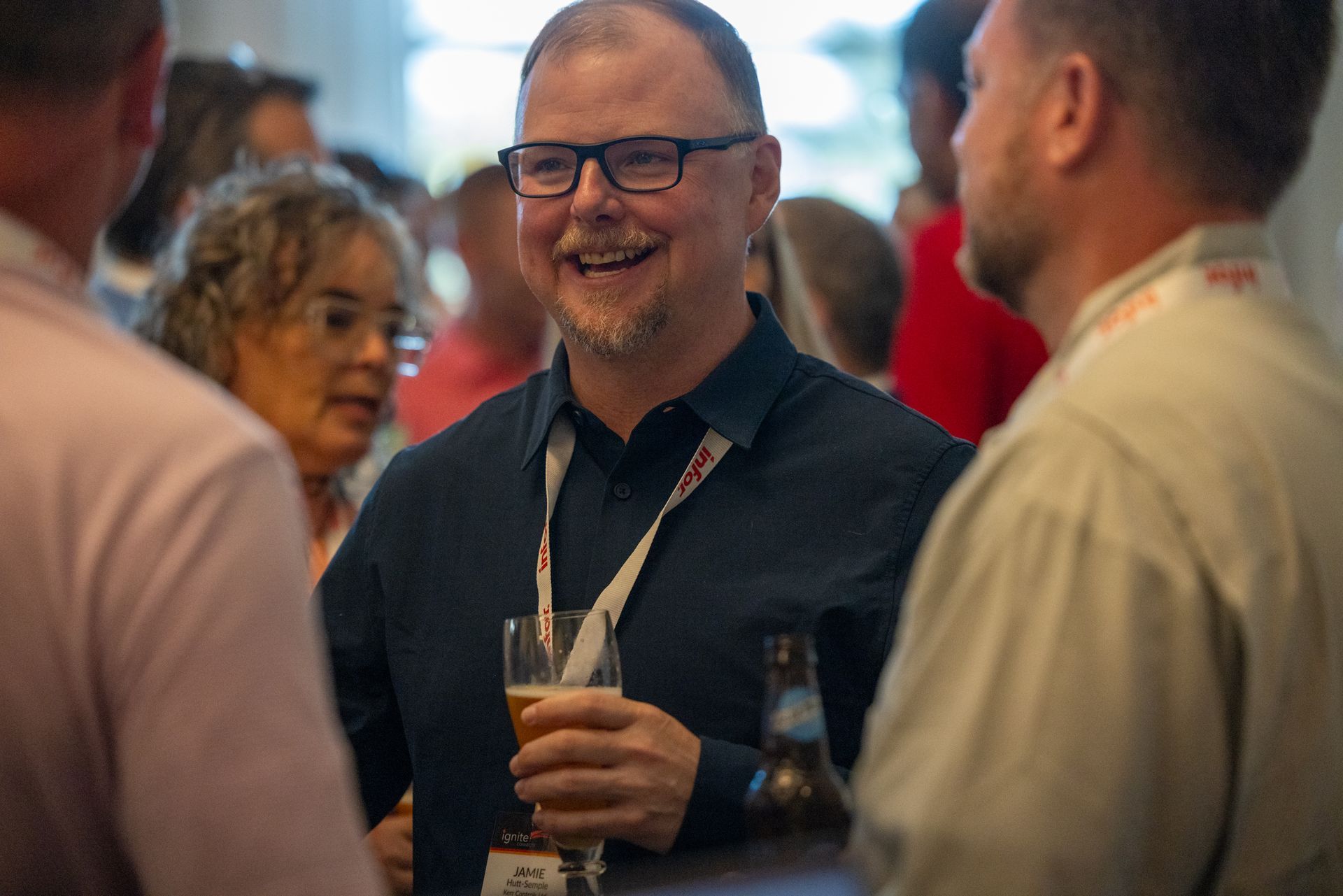 A man (Jamie Hutt-Semple) wearing a white lanyard over a dark collared shirt, smiles at another man.