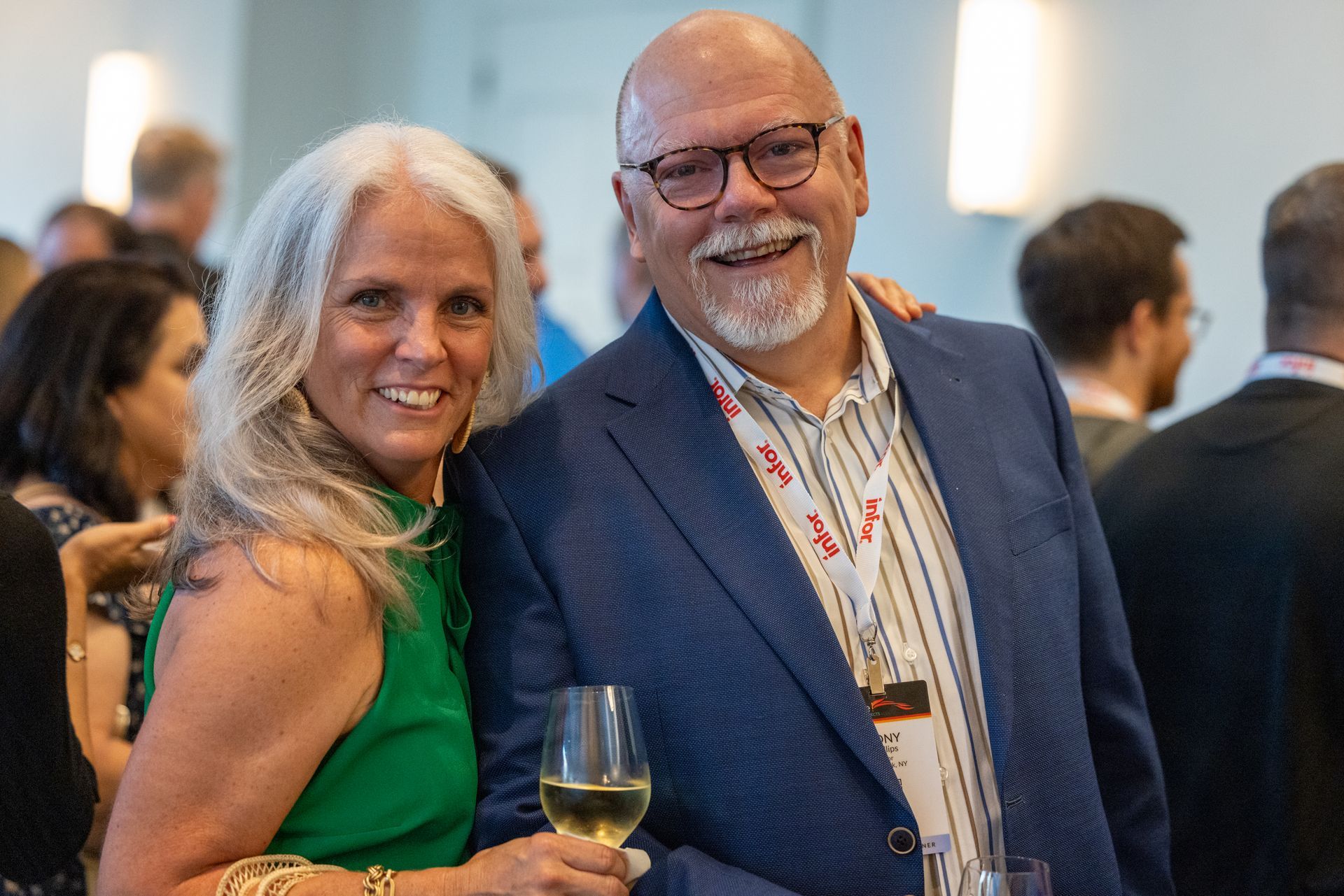 A woman with long gray hair wearing a green dress and a bald man with a gray goatee wearing a blue suit jacket and striped shirt smile at the camera