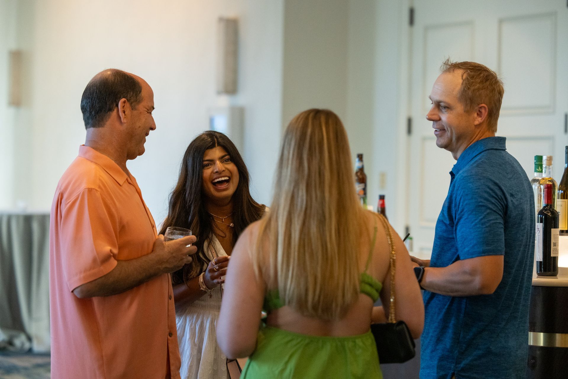 A woman with long dark hair smiles at the camera. A man on her right wears a blue shirt looks at her and smiles, a man on her left wears a pink shirt, also looks at her smiling. A woman with long blond hair faces away from the camera
