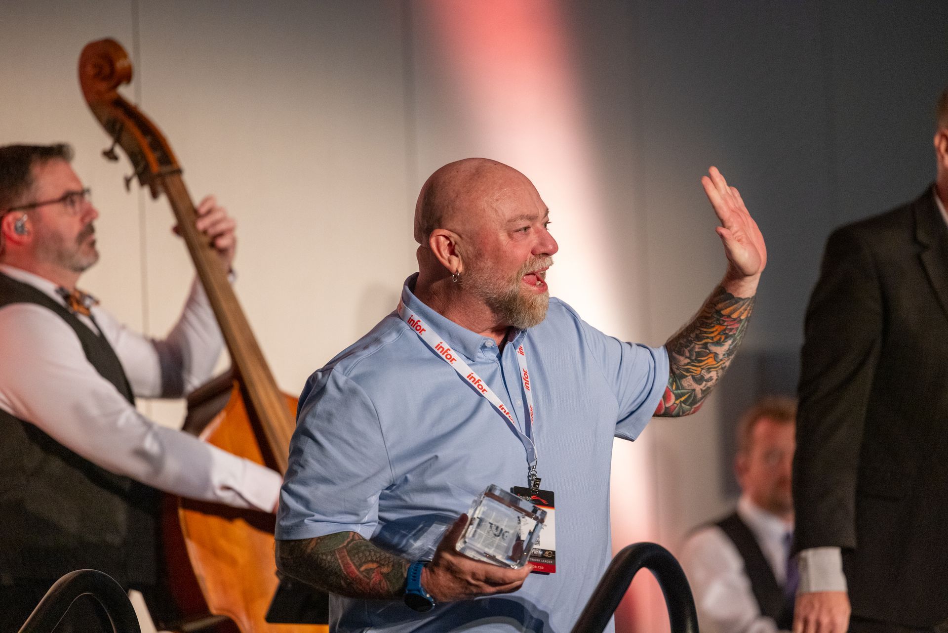 A cello player in the background, a bald man holding a crystal award waves to the right.
