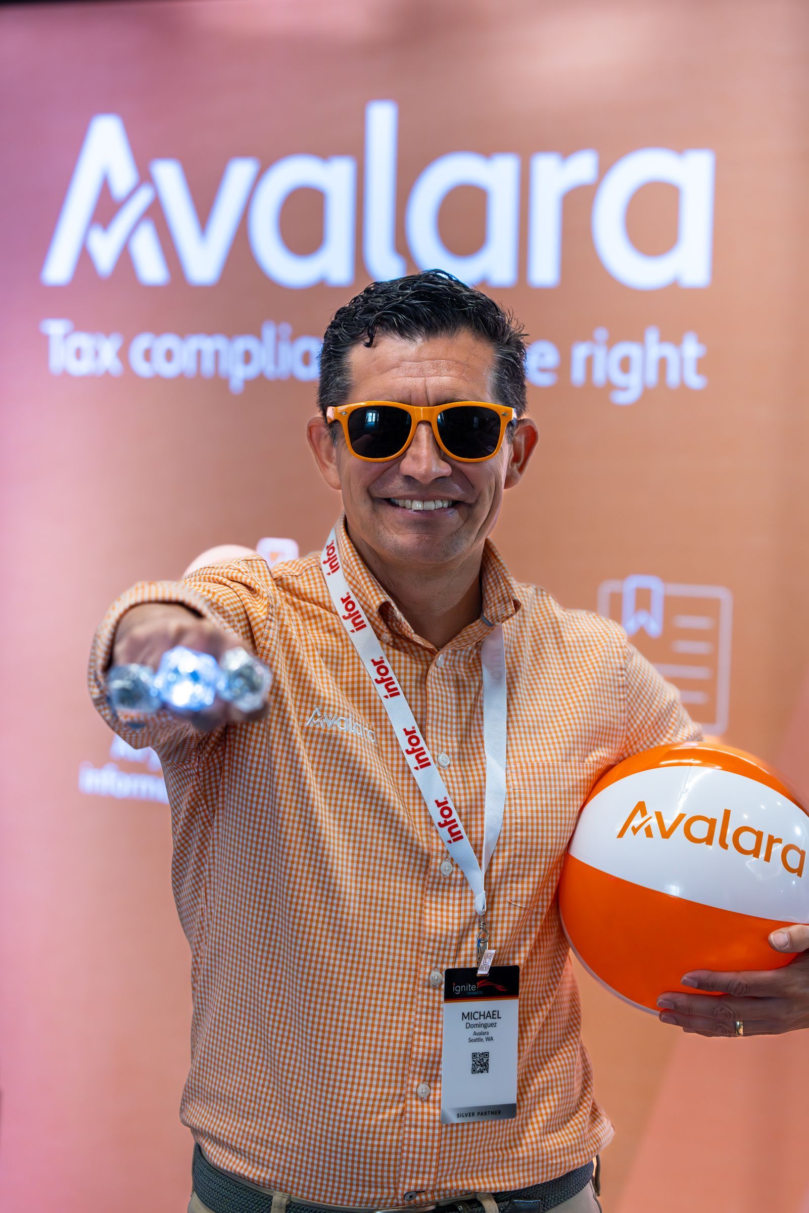 A man in an orange shirt offers up some swag to the viewer. Behind him is a sign for Avalara