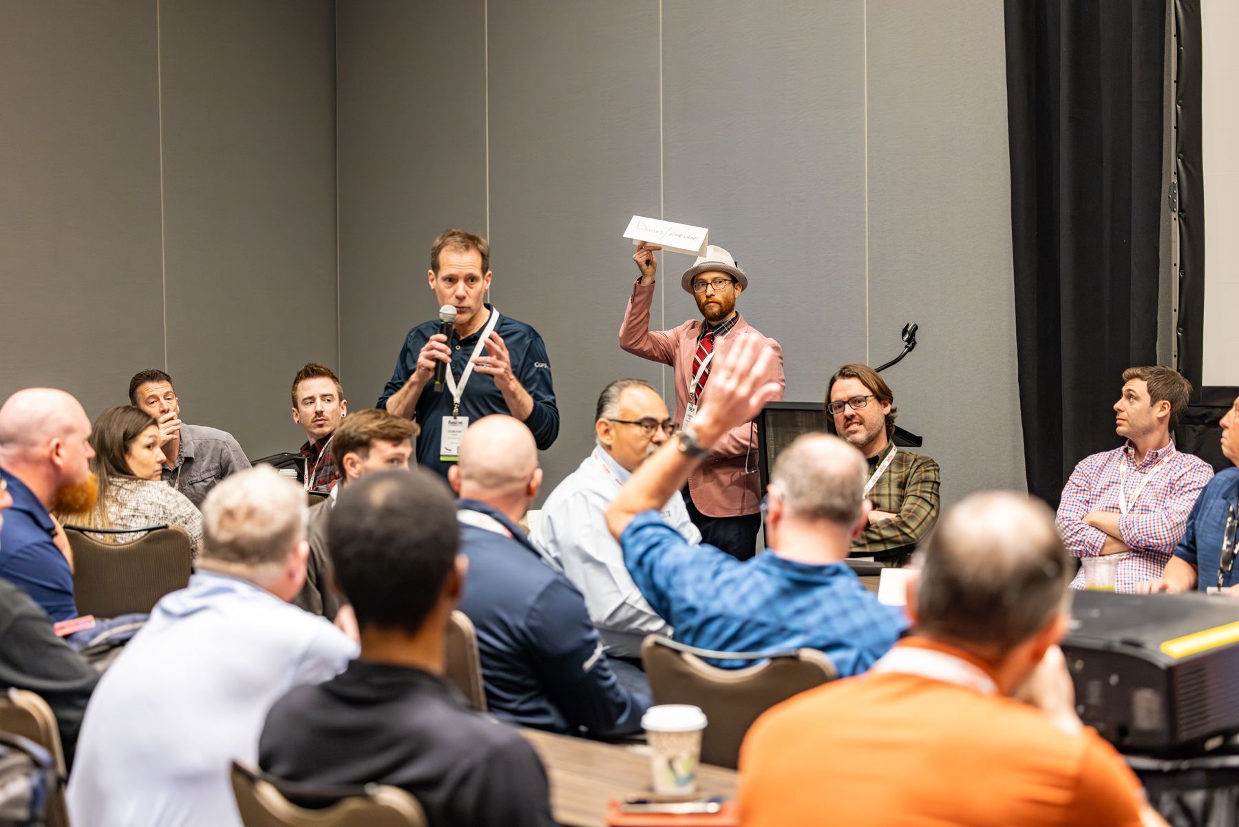 A group of people in a conference room. Two men are standing in front, one holding a paper and gesturing.