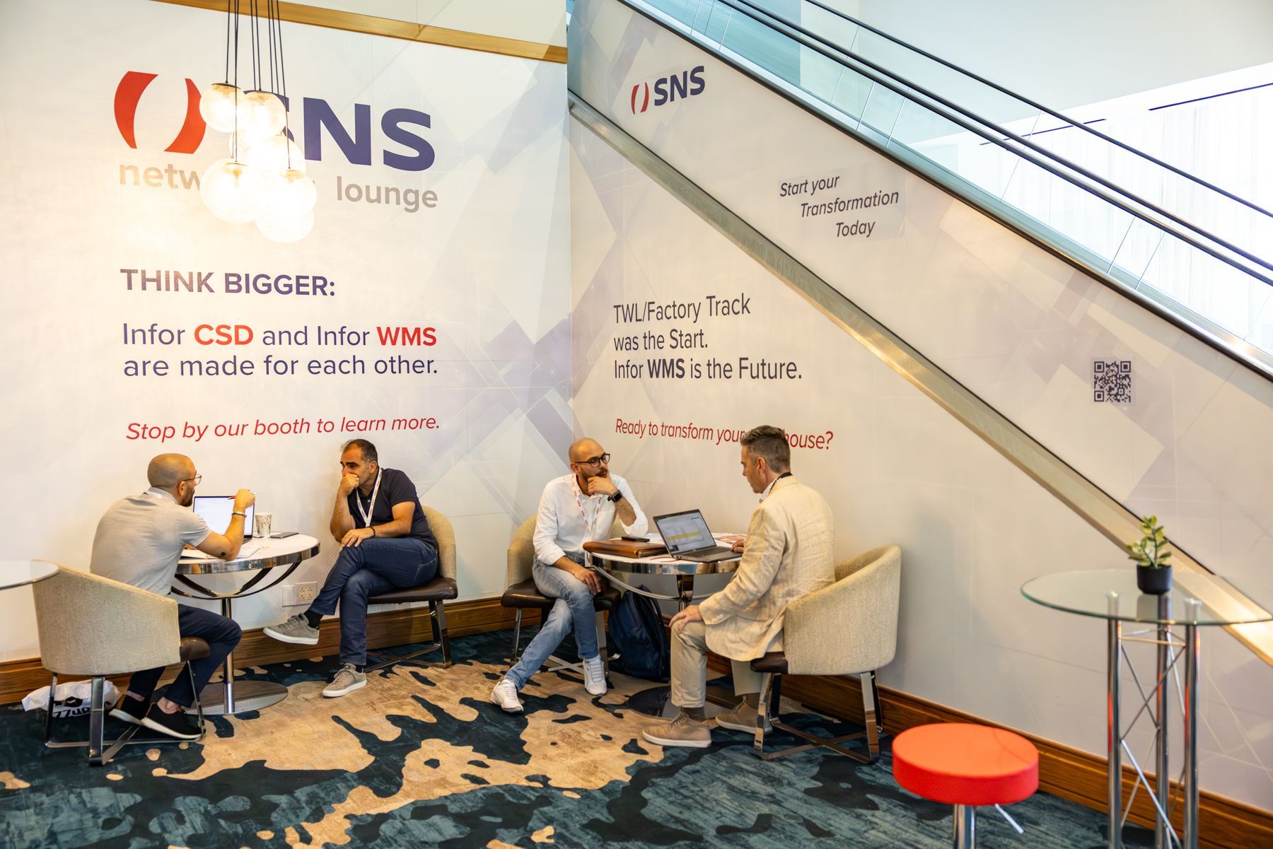 Four people at a networking lounge, using laptops. The lounge has a patterned wall and a red and white chair.