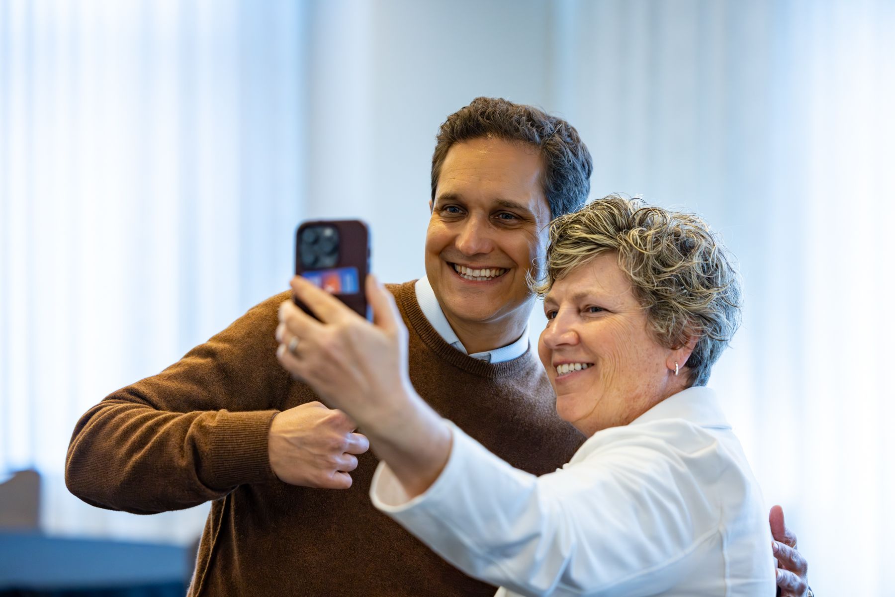 Man in brown sweater smiles while woman takes selfie. Interior, bright light.
