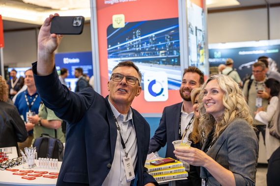 A man in a blazer takes a selfie with colleagues at a trade show exhibit, while a woman beside him holds a drink.