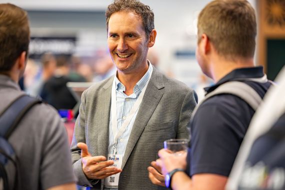 A man in a grey blazer smiles while talking to two people at a conference, holding a name tag and a glass.