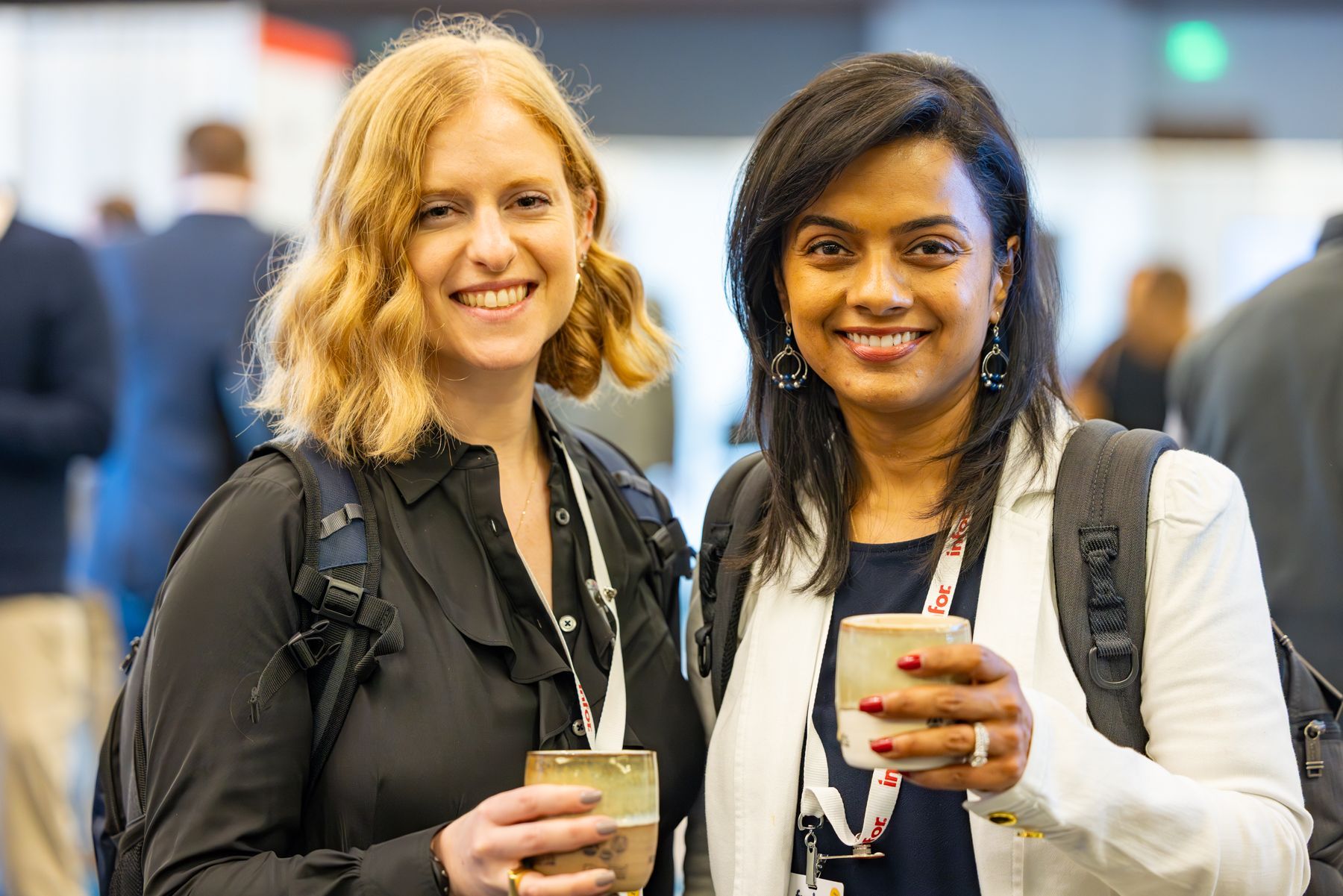 Two women smiling, holding drinks, wearing lanyards, and standing in a conference setting.