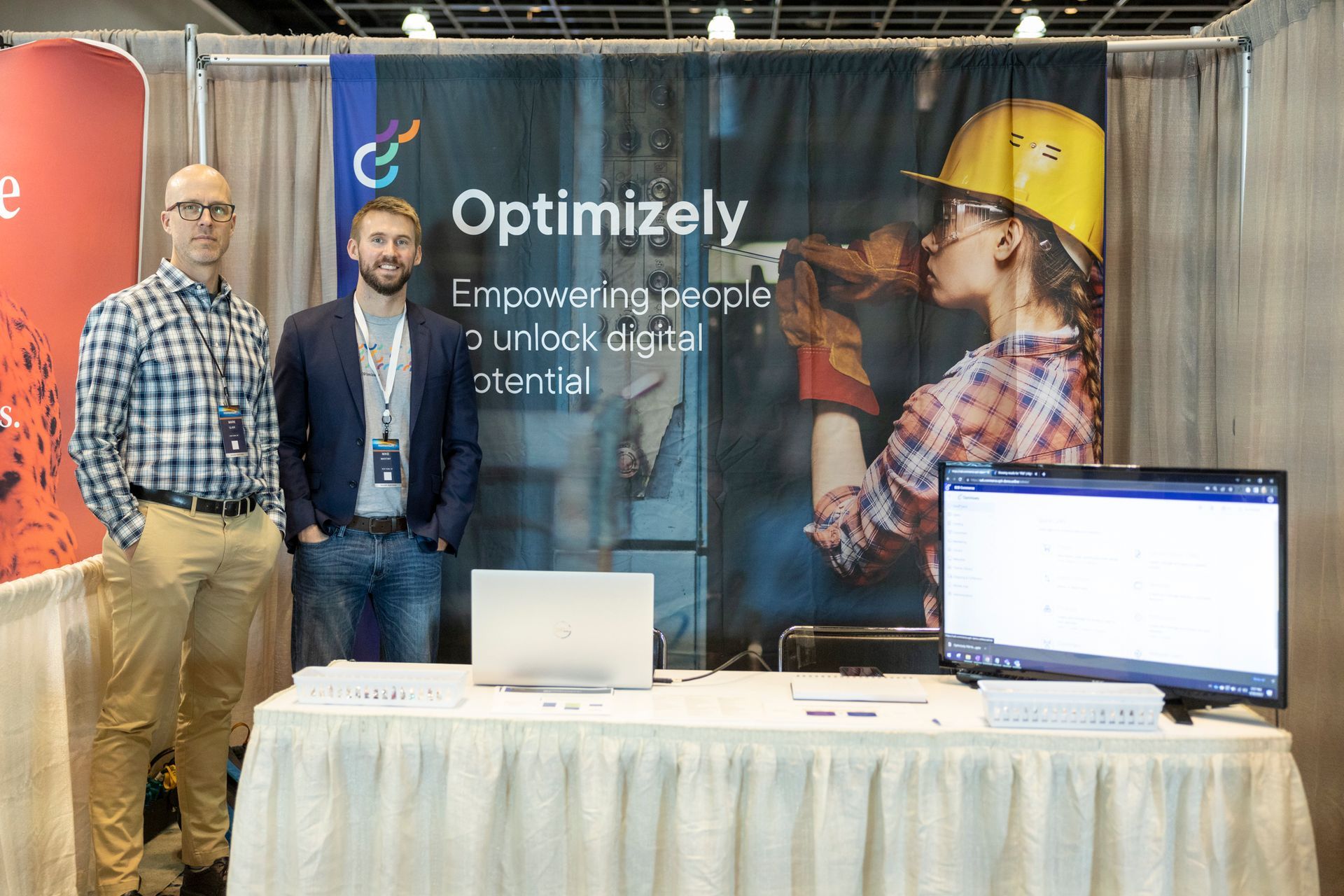 Optimizely booth featuring two men standing to the left of a white skirted table. Behind them a mural reads Optimizely - Empowering People to unlock digital potential