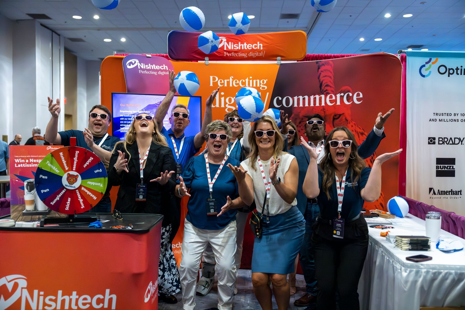 Nishtech booth featuring nine very happy men and women, all wearing sunglasses, tossing blue and white balls in the air.