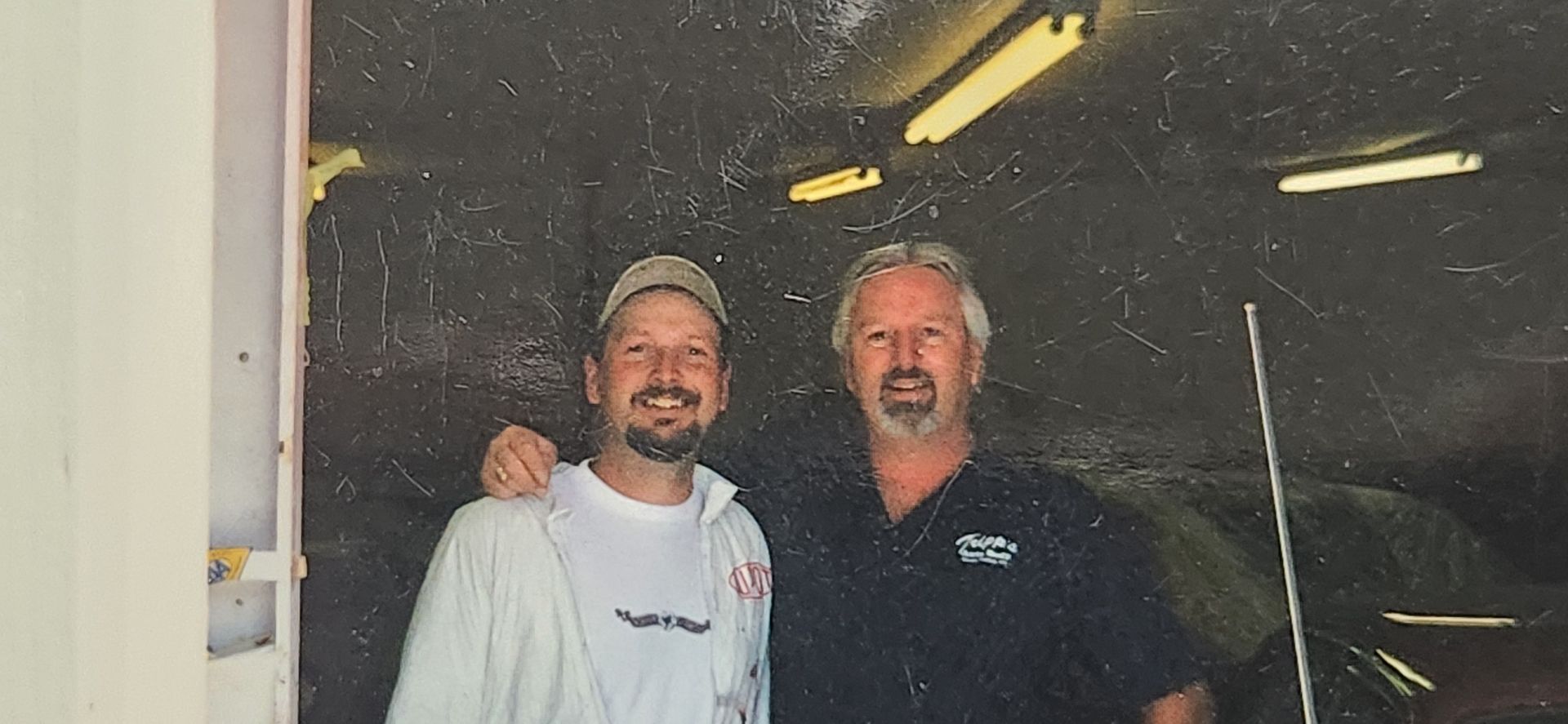 Two smiling men stand together in a workshop doorway, one in a white shirt and cap, the other in a dark shirt. | Tripp's Auto Body