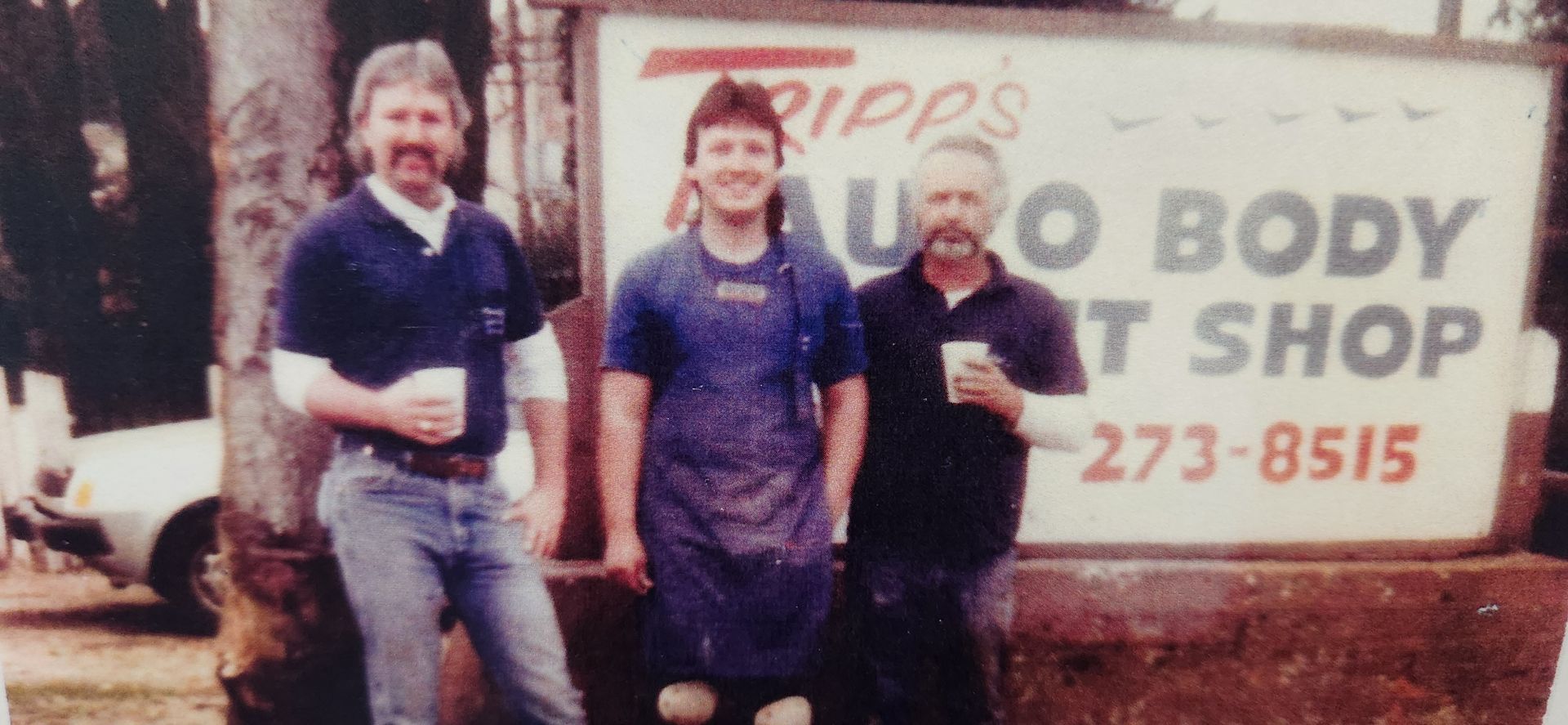 Three people standing in front of a sign for an auto body shop, smiling and holding drinks | Tripp's Auto Body