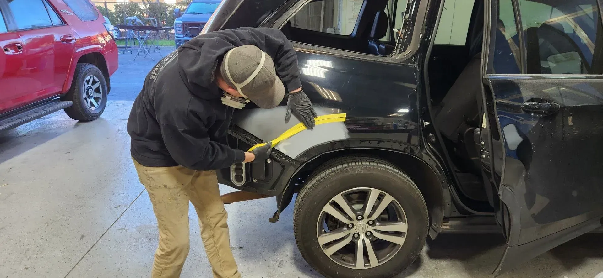 Person working on the rear wheel area of a black SUV in a garage, with a red car nearby. | Tripp's Auto Body