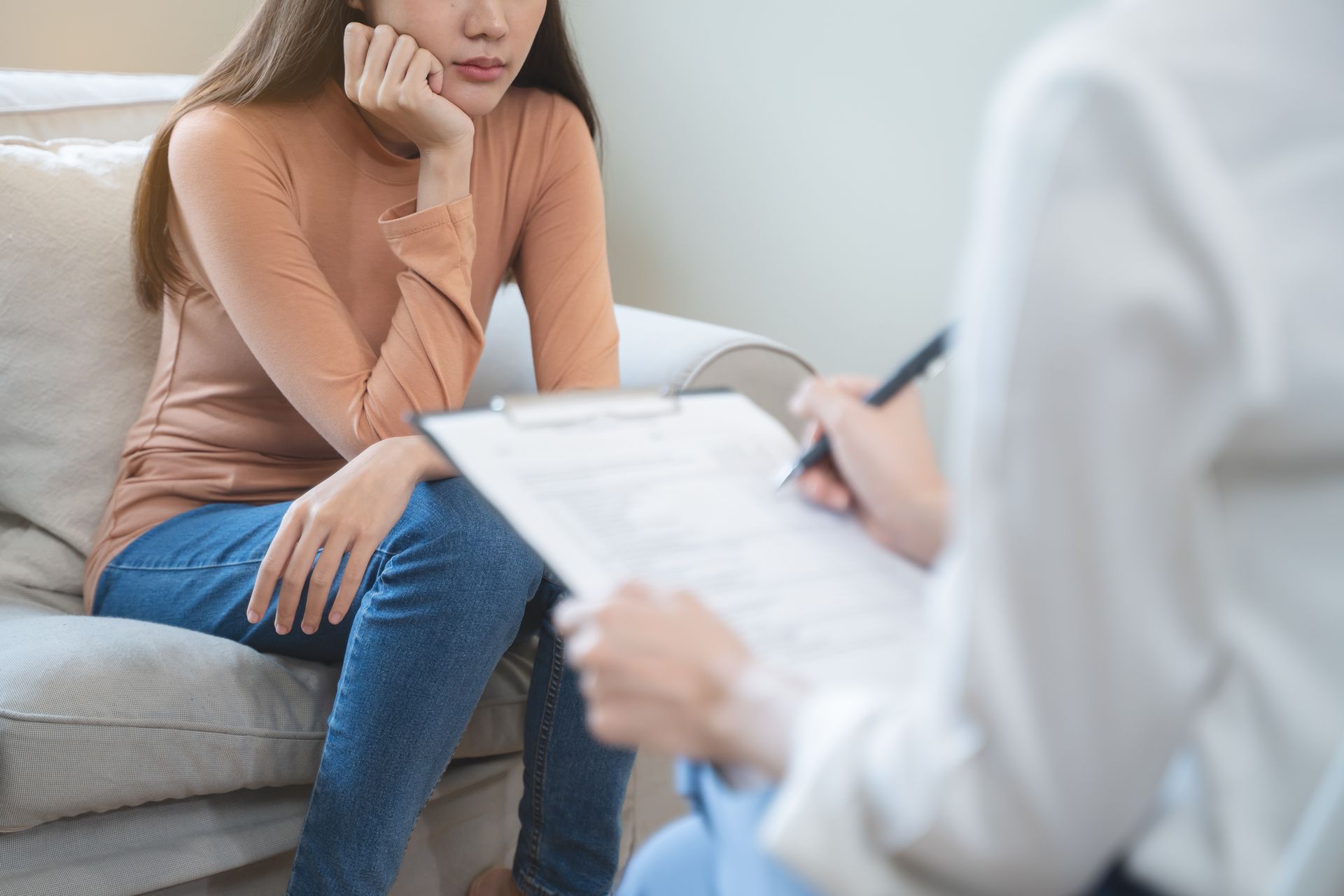 Woman in therapy session, sitting on a couch, talking to a person holding a clipboard and pen.