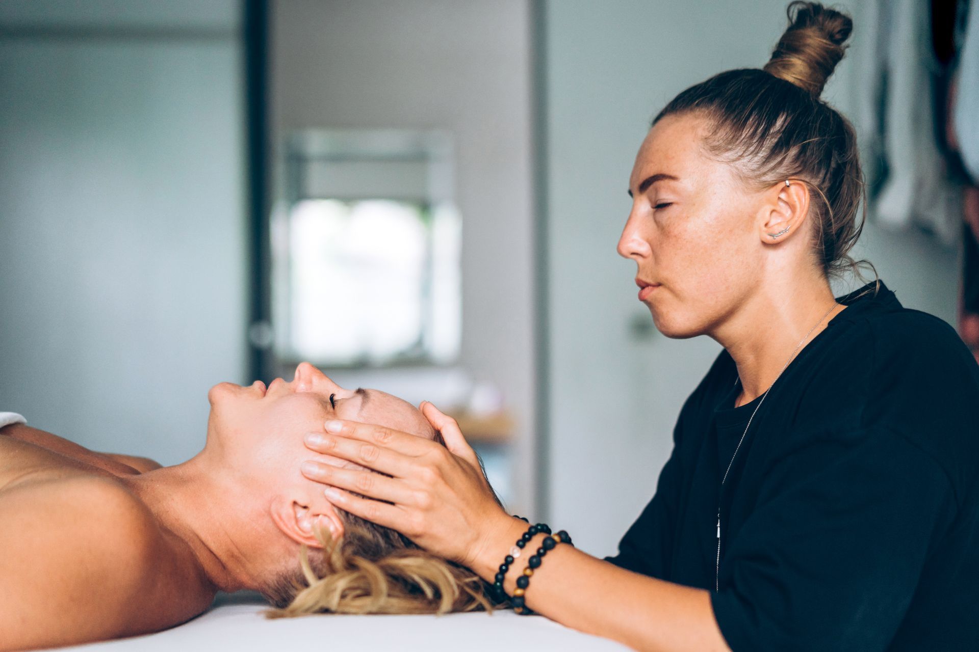 Woman performing a facial massage on another woman lying on a massage table. Inside, light-colored room.