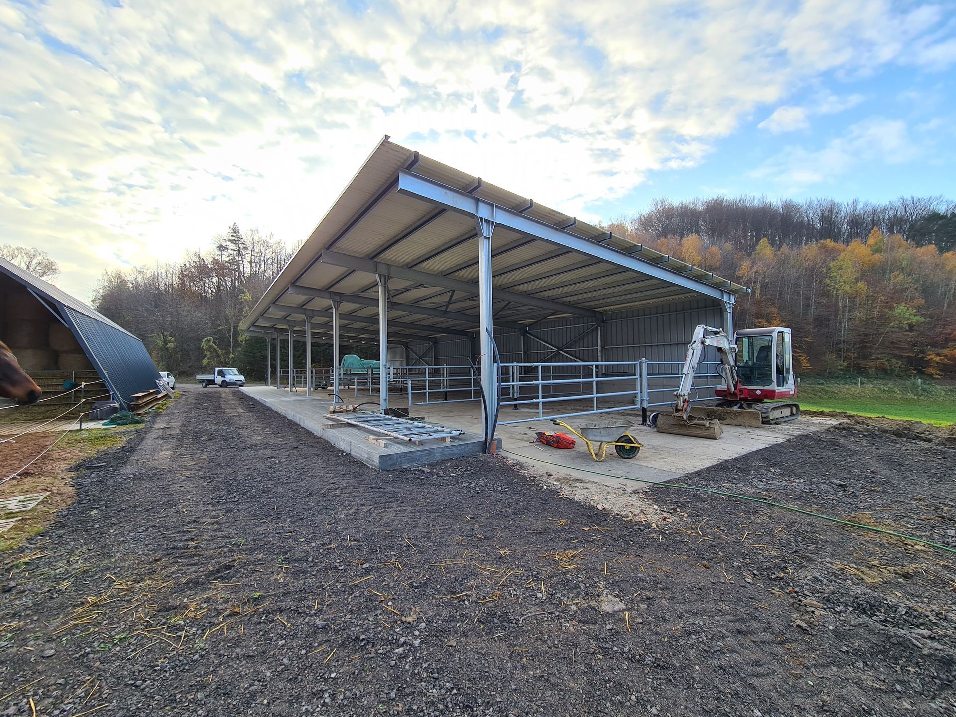 Scheunenkonstruktion mit Metallrahmen. Kiesboden, kleiner Bagger und blauer Himmel mit Wolken im Hintergrund.