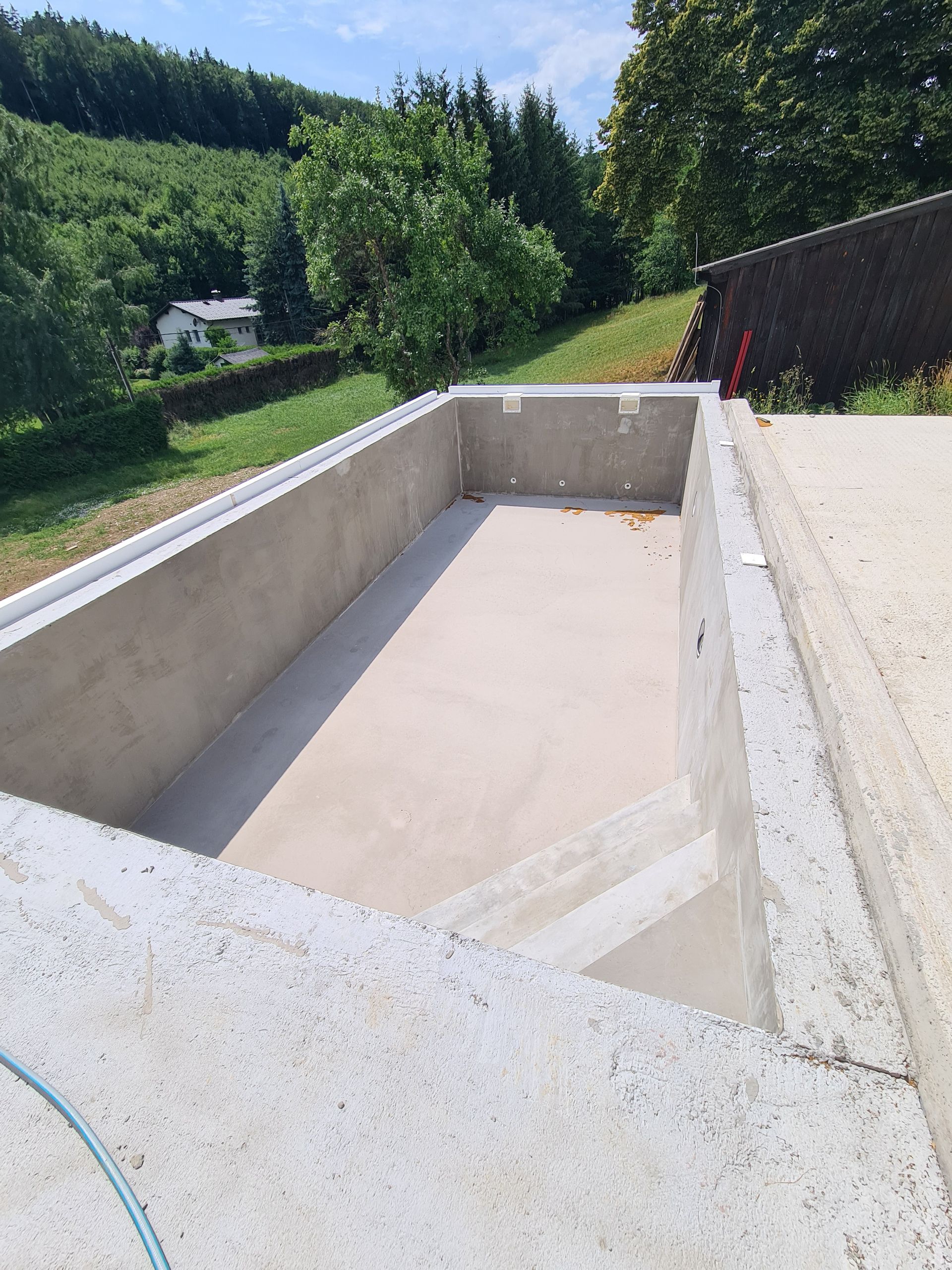 Leeres Betonschwimmbecken im Bau, im Freien mit Bäumen im Hintergrund.