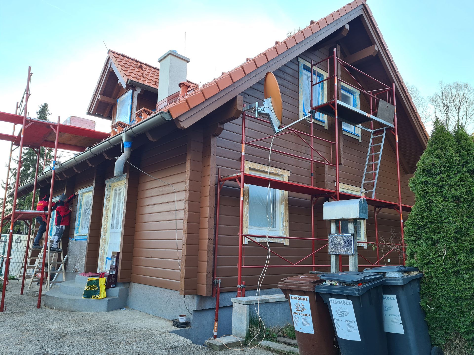 Braunes zweistöckiges Haus im Umbau mit Gerüst. Hellbraunes Dach, braune Außenverkleidung und blaue Fensterrahmen.
