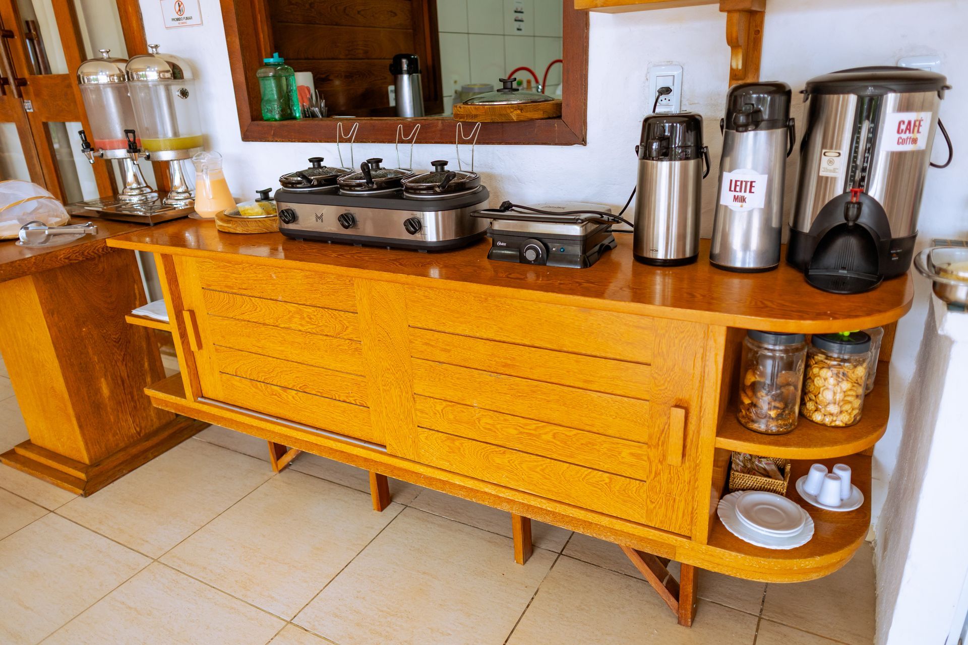 Aparador de madeira com portas de correr, dois fogões pequenos e dispensadores de café/chá na parte superior; prateleiras para guardar potes e pratos.