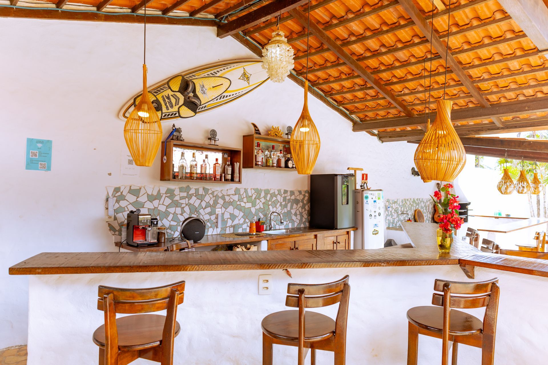 Bar ao ar livre com balcão de madeira, banquetas e luminárias pendentes. Prateleira com garrafas e uma prancha de surfe acima.