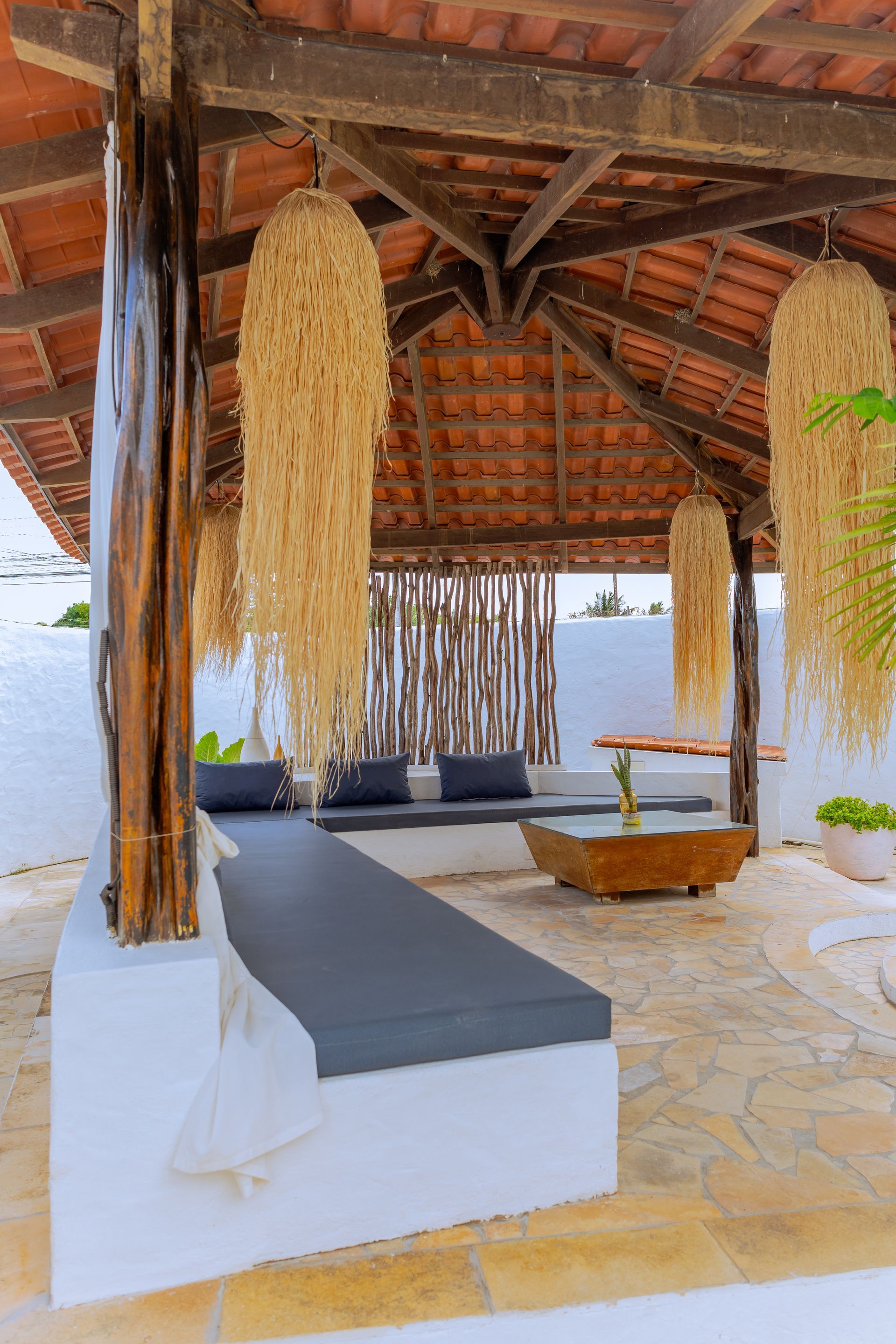 Área de estar externa sob um pergolado de madeira. Paredes brancas, almofadas cinza, decoração com palha pendurada.