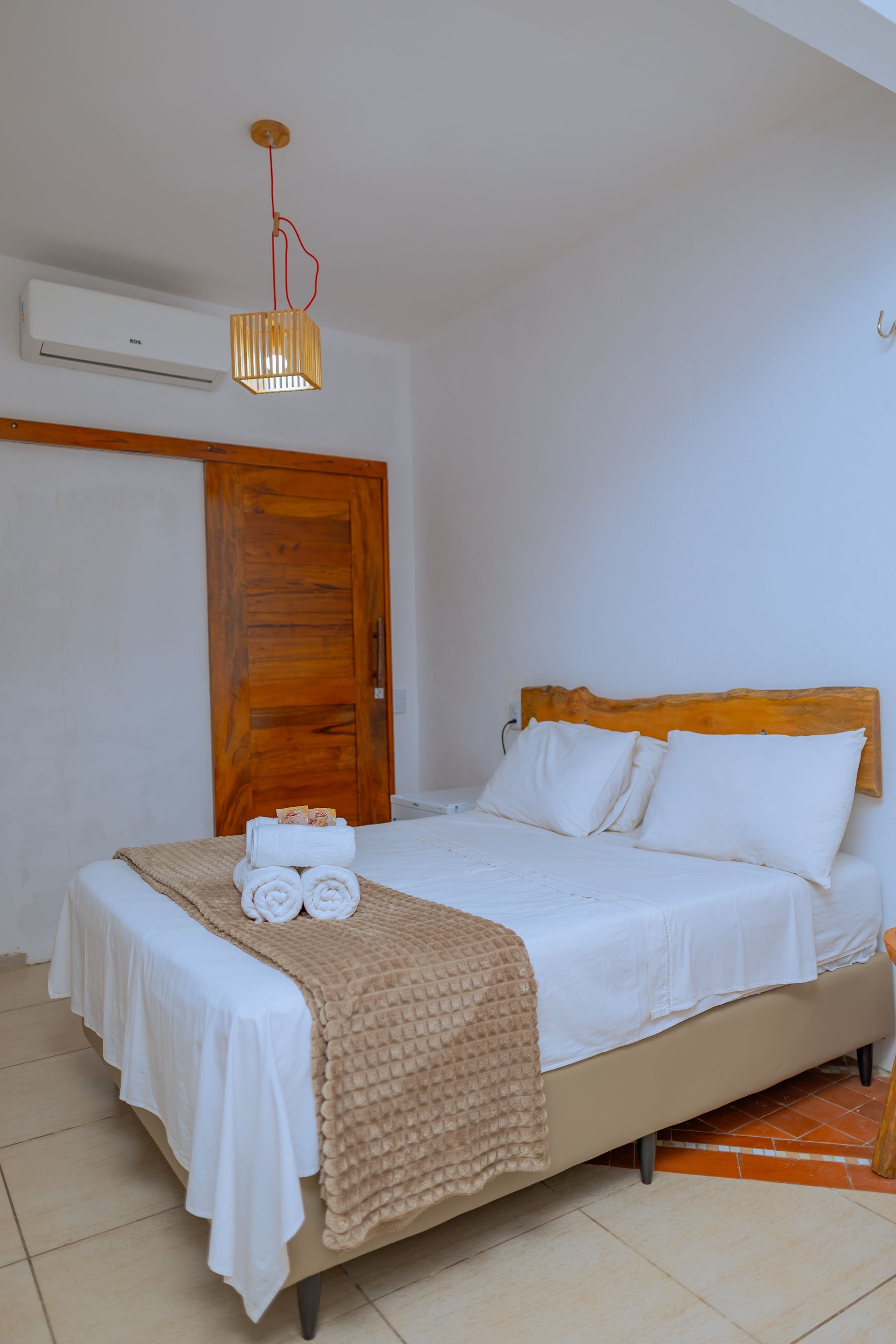 Quarto com roupa de cama branca, cabeceira marrom, manta bege e porta de madeira.