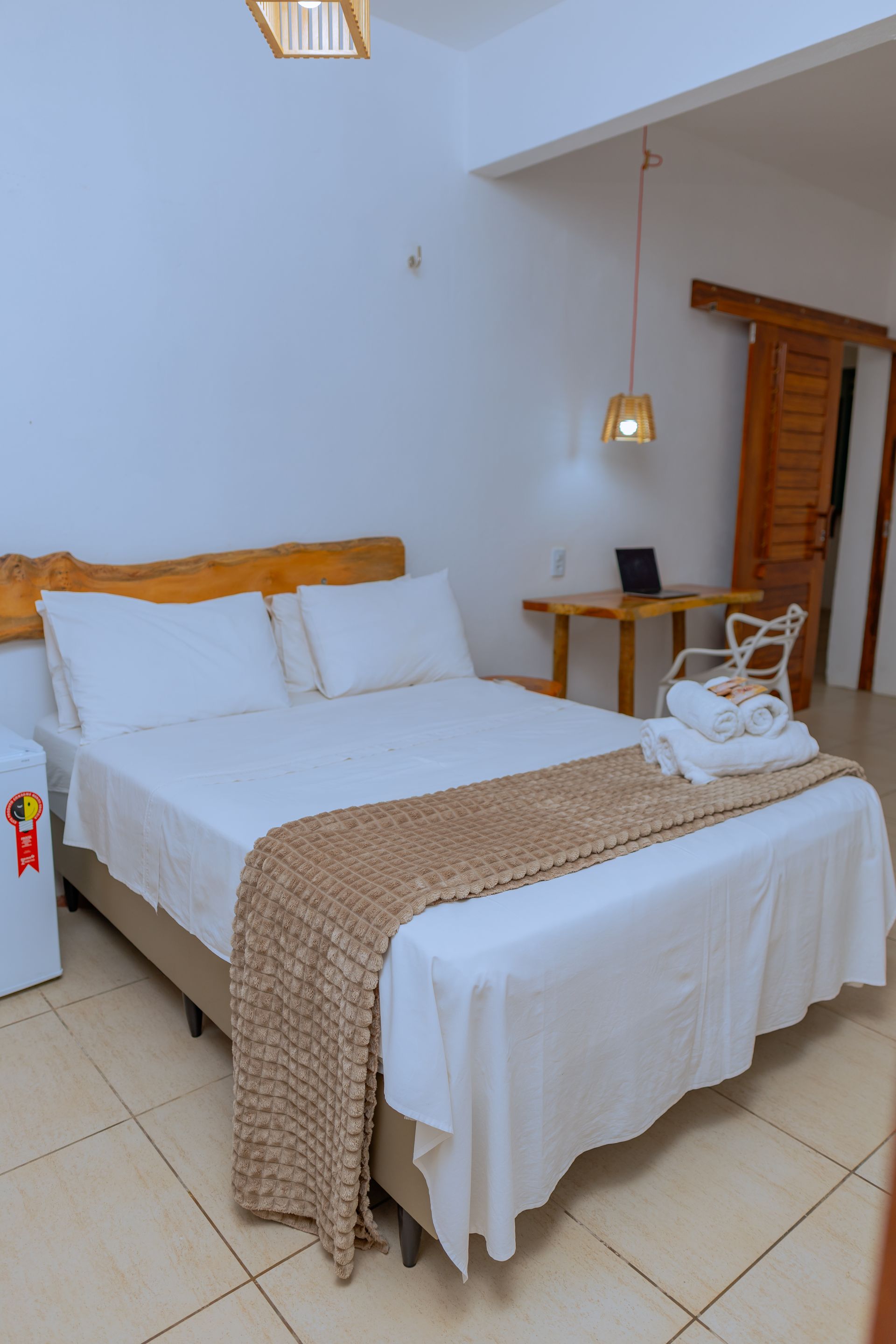 Quarto com cama, lençóis brancos, cabeceira de madeira, frigobar, escrivaninha e porta aberta.