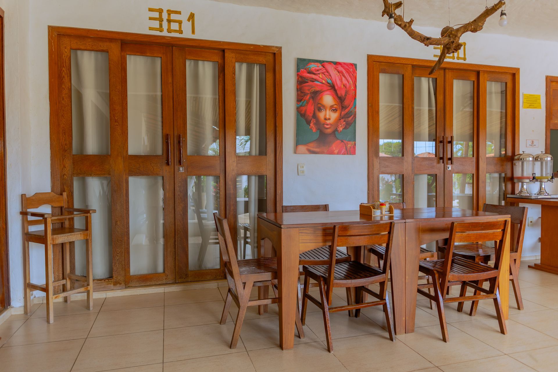Sala de jantar interna com portas de madeira, mesa, cadeiras e obra de arte representando uma pessoa com um turbante vermelho.