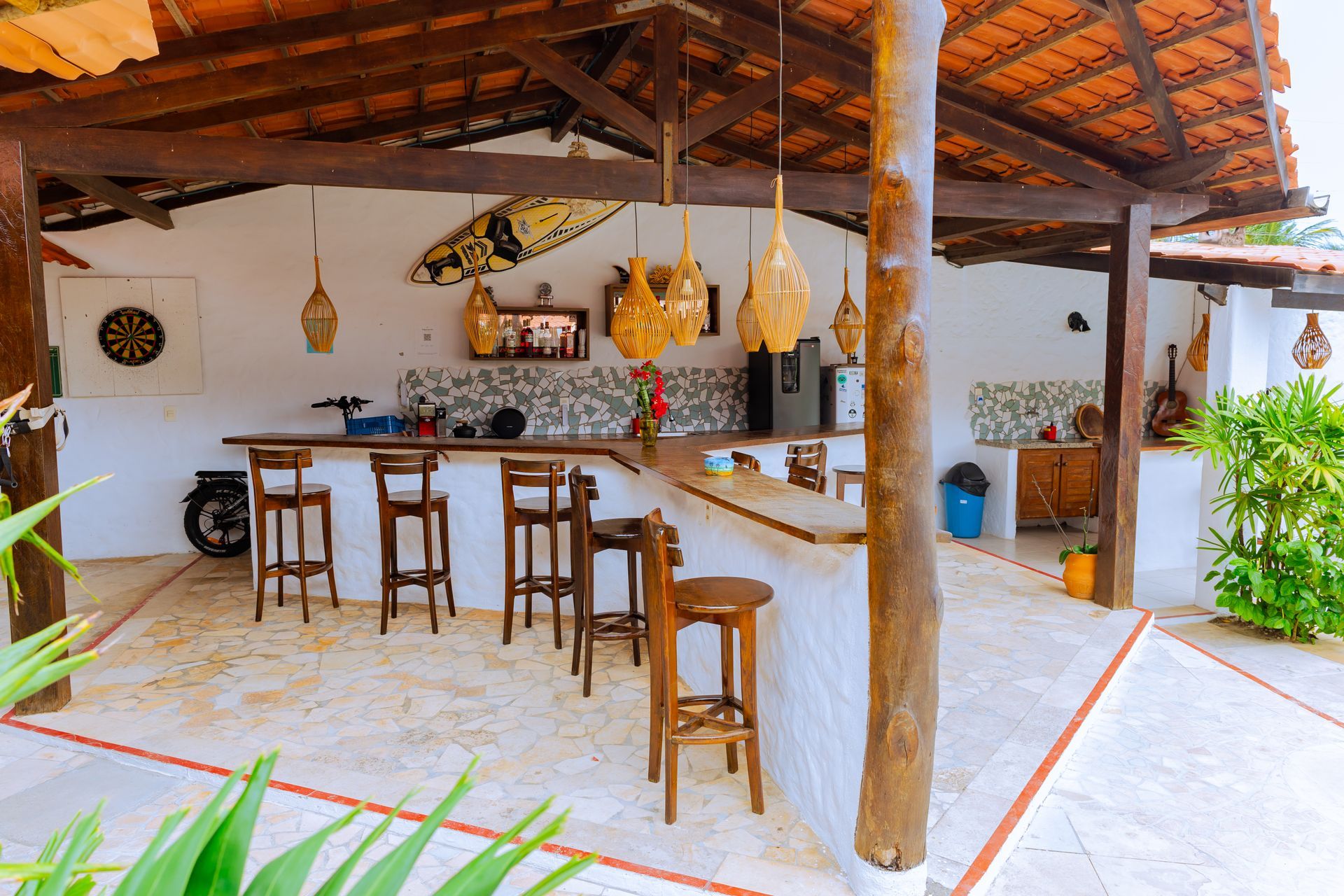 Bar ao ar livre com banquetas, iluminação suspensa e paredes brancas sob um telhado de madeira.