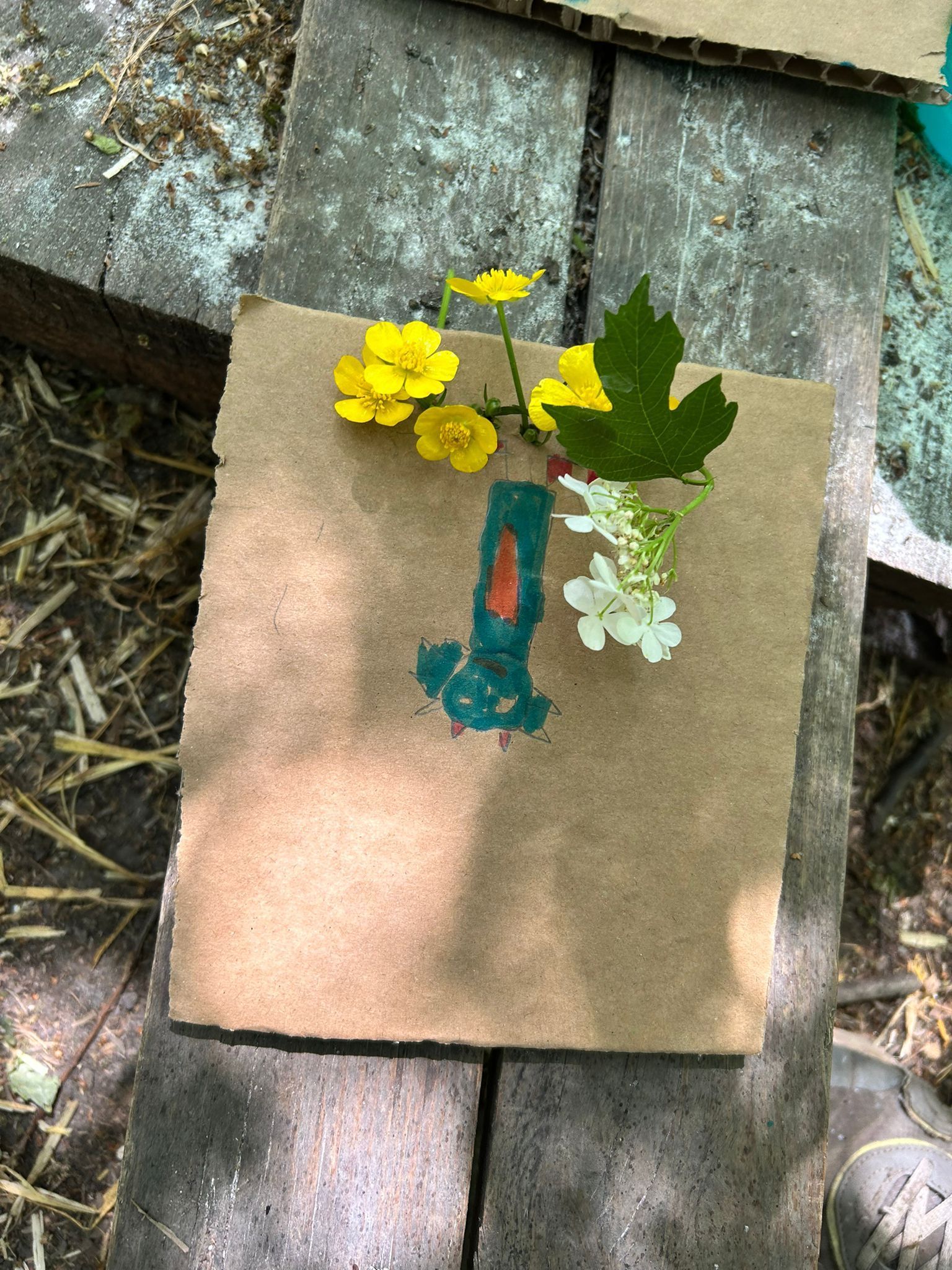 Cardboard with a painted vase holding yellow flowers, white flowers, and a green leaf.