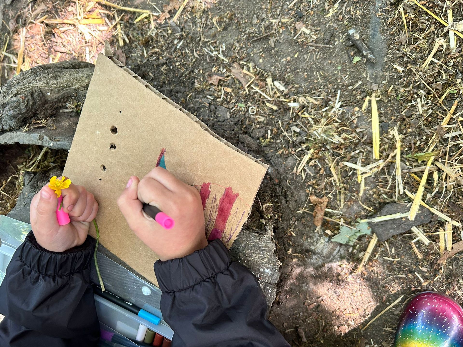 Child drawing on cardboard outdoors, holding a flower.
