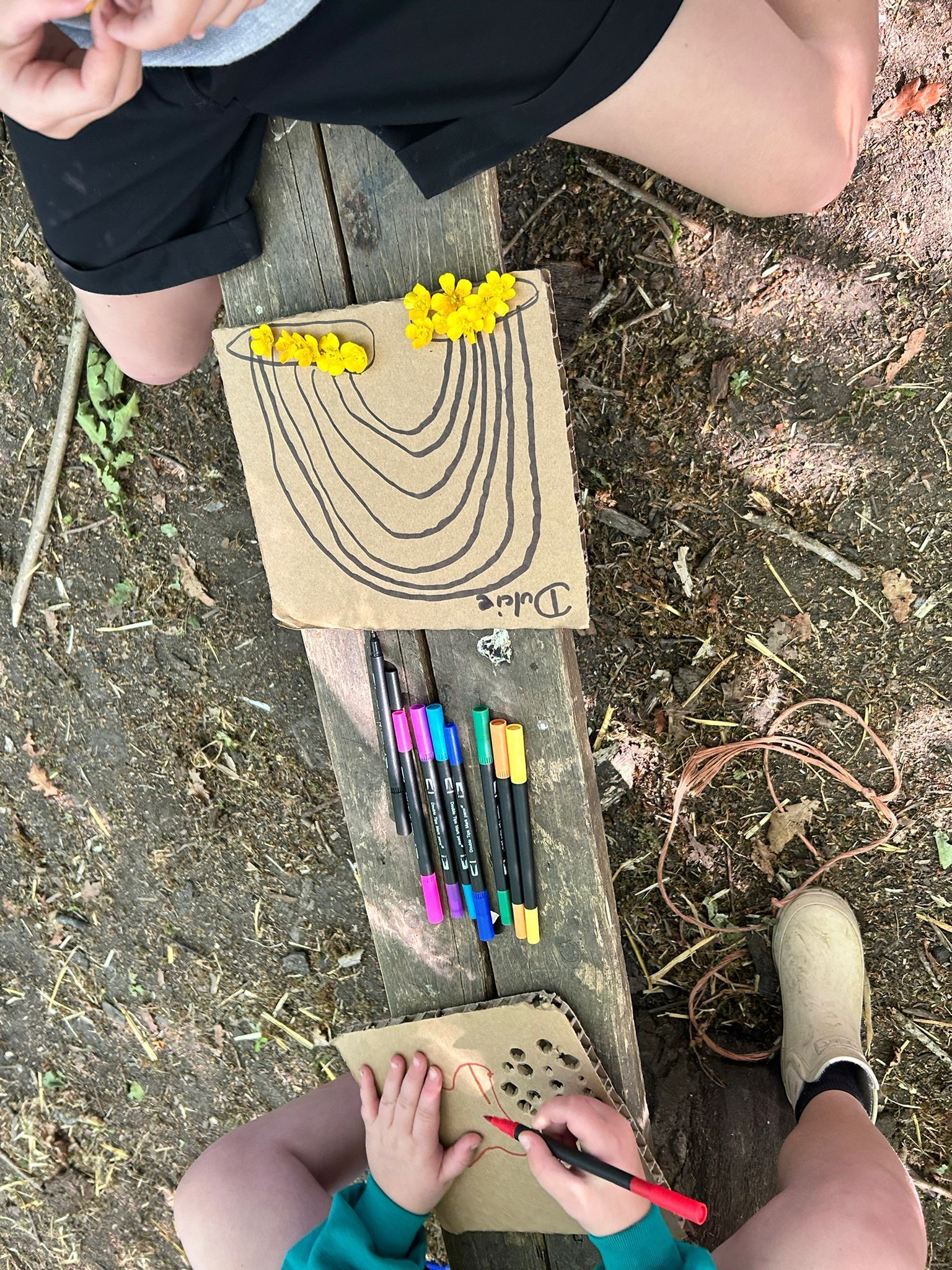 Two people drawing on cardboard with markers outdoors. One person has a finished drawing of a rainbow.