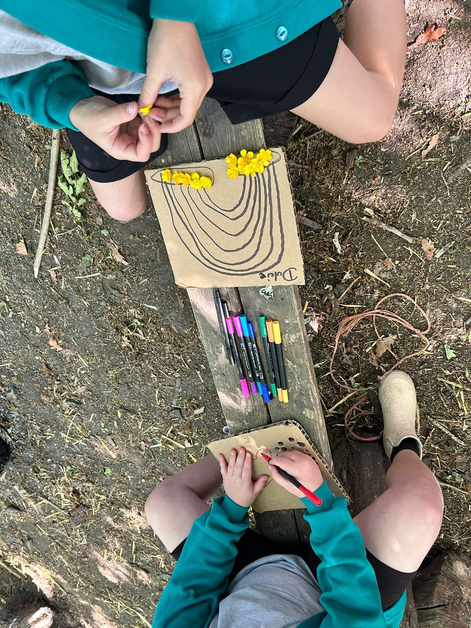 Two children seated outdoors draw on paper and play with small yellow objects.
