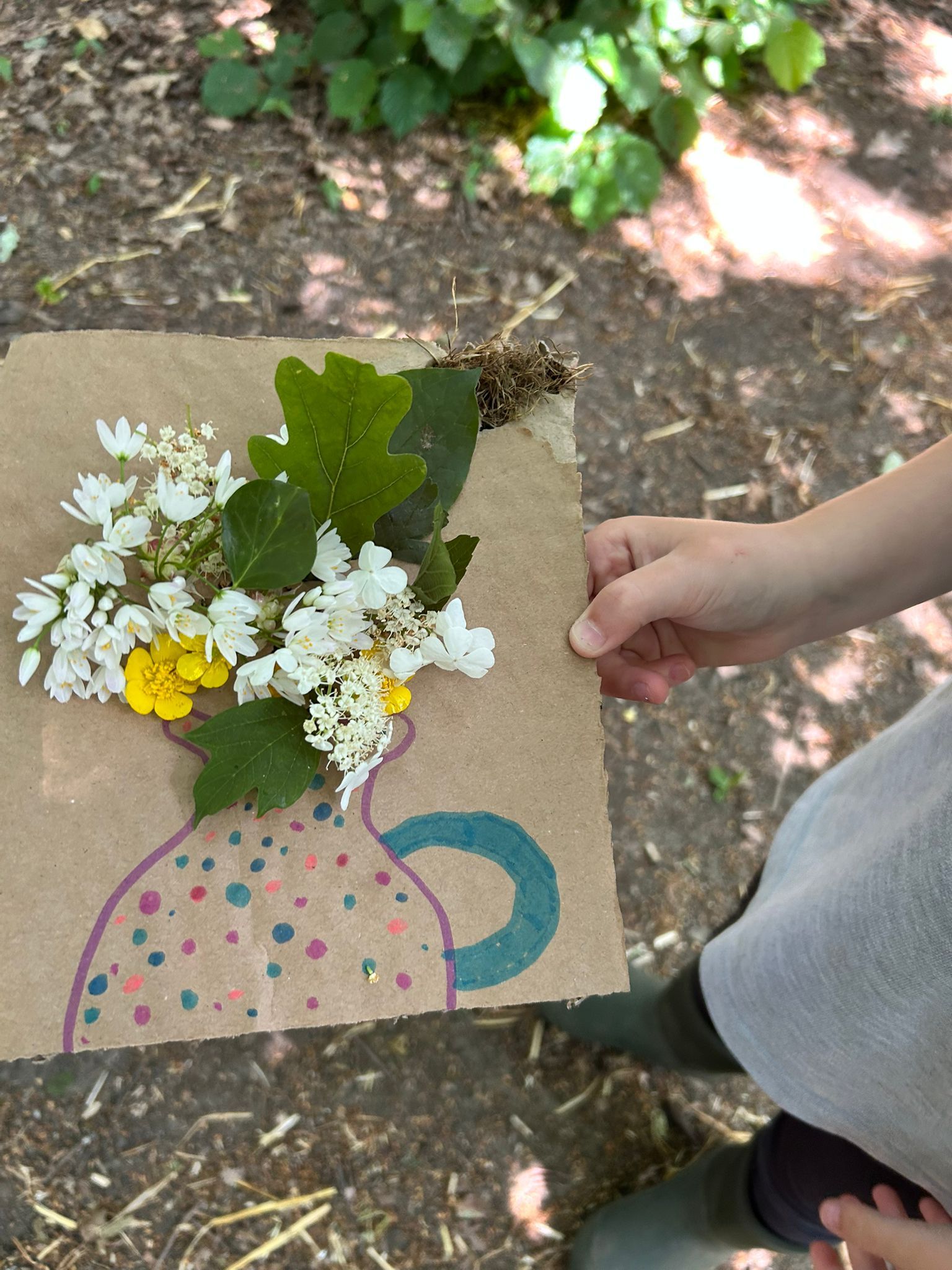 Child's hand holding a craft of flowers and leaves arranged above a drawing of a vase on brown paper.