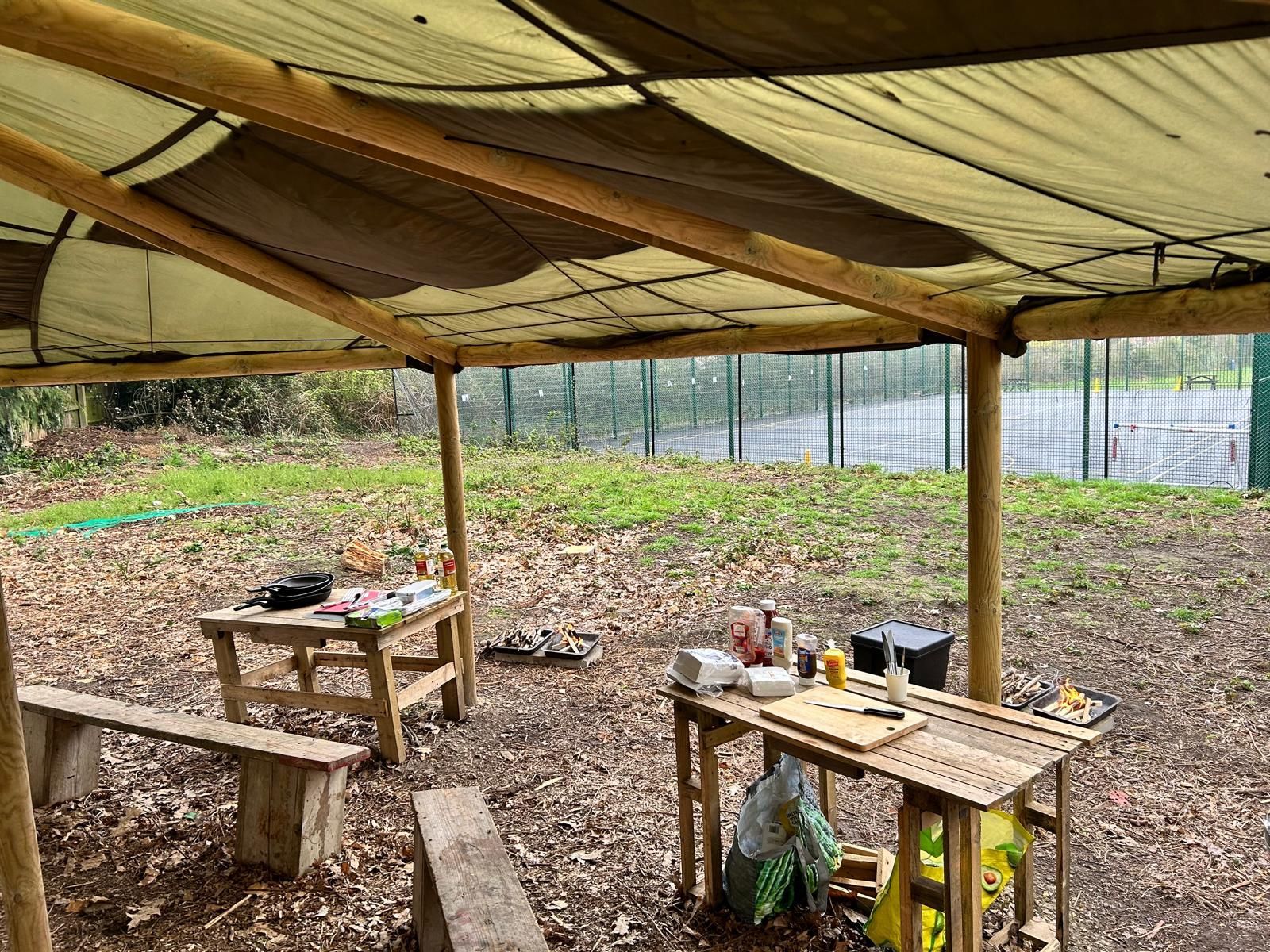 A wooden table and benches under a canopy in the woods.