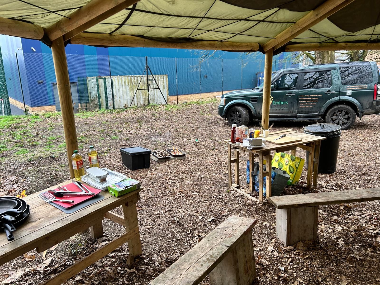 A wooden picnic table and benches under a canopy with a car parked in the background.