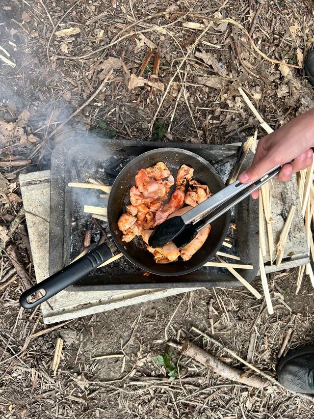 A person is cooking food in a frying pan on an outdoor fire.