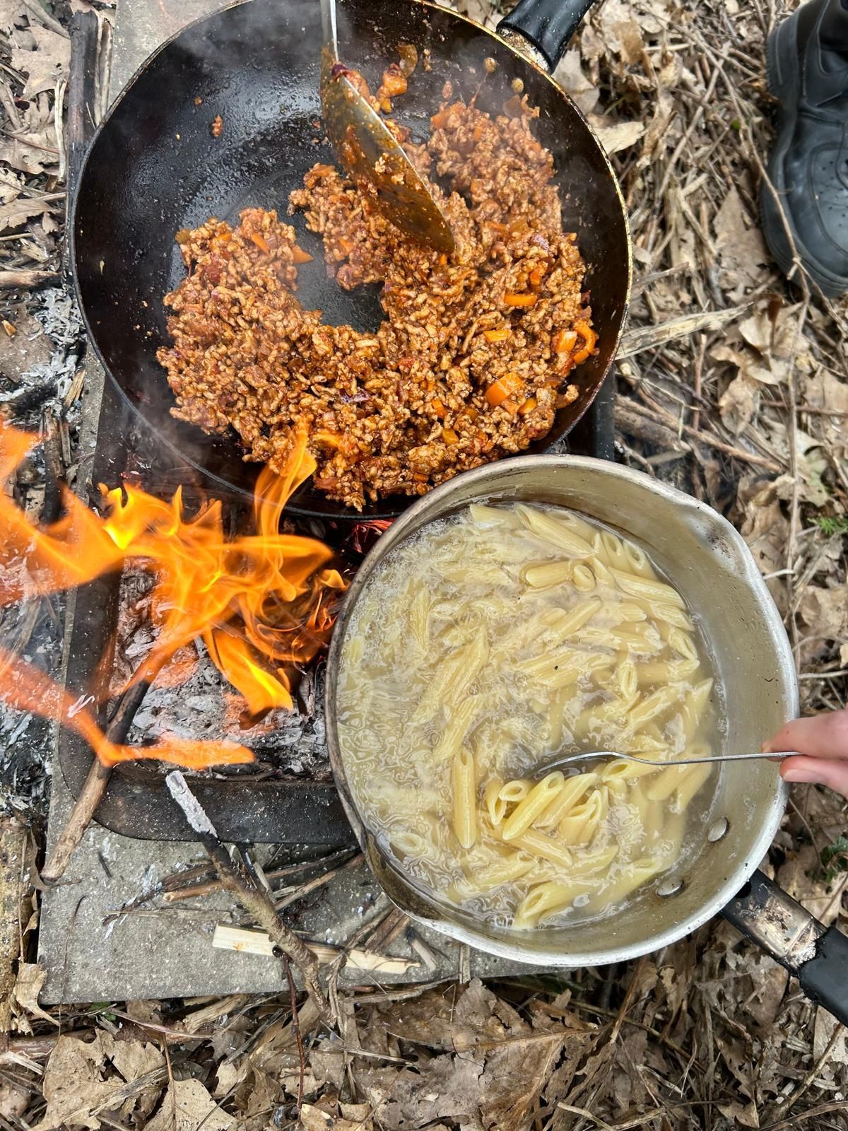A person is stirring a pot of pasta next to a pan of food on a fire.
