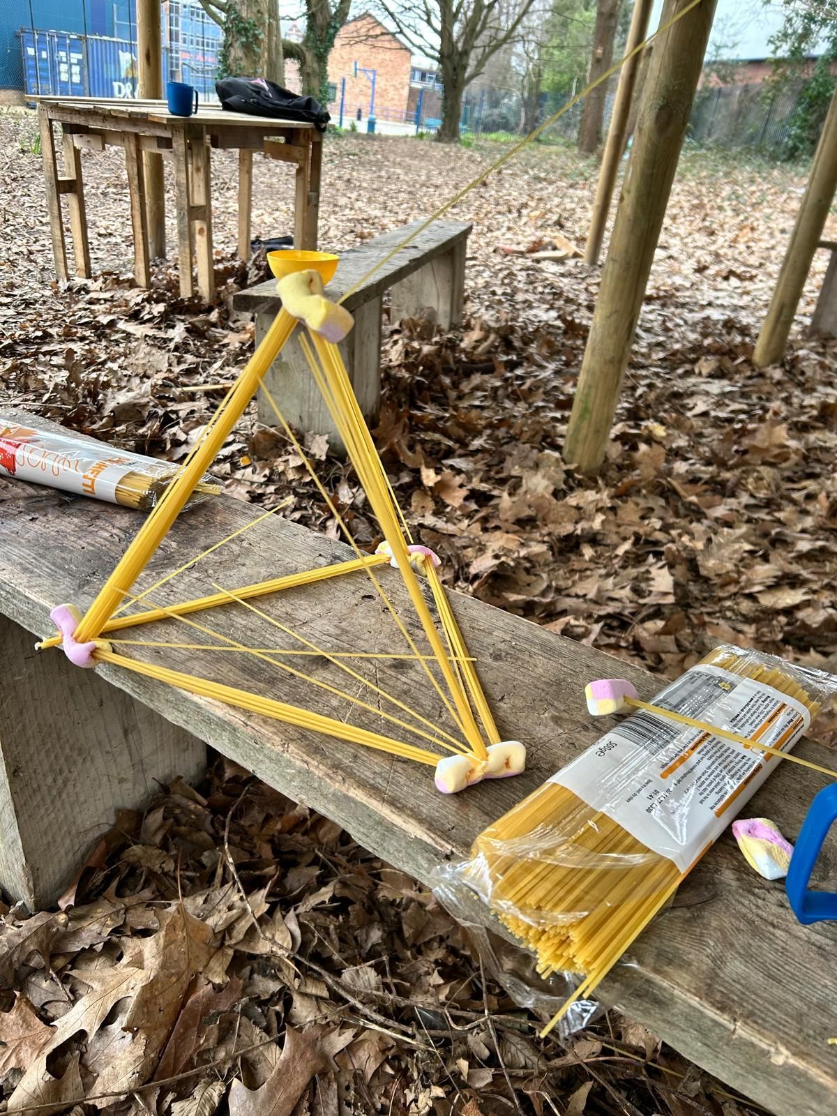 A triangle made out of spaghetti and marshmallows is sitting on a wooden bench.
