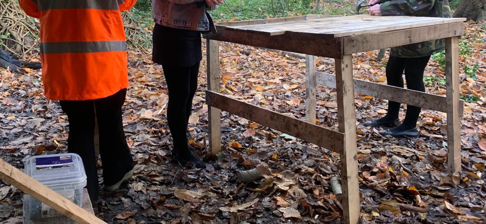 A group of people are standing around a wooden table in the woods.